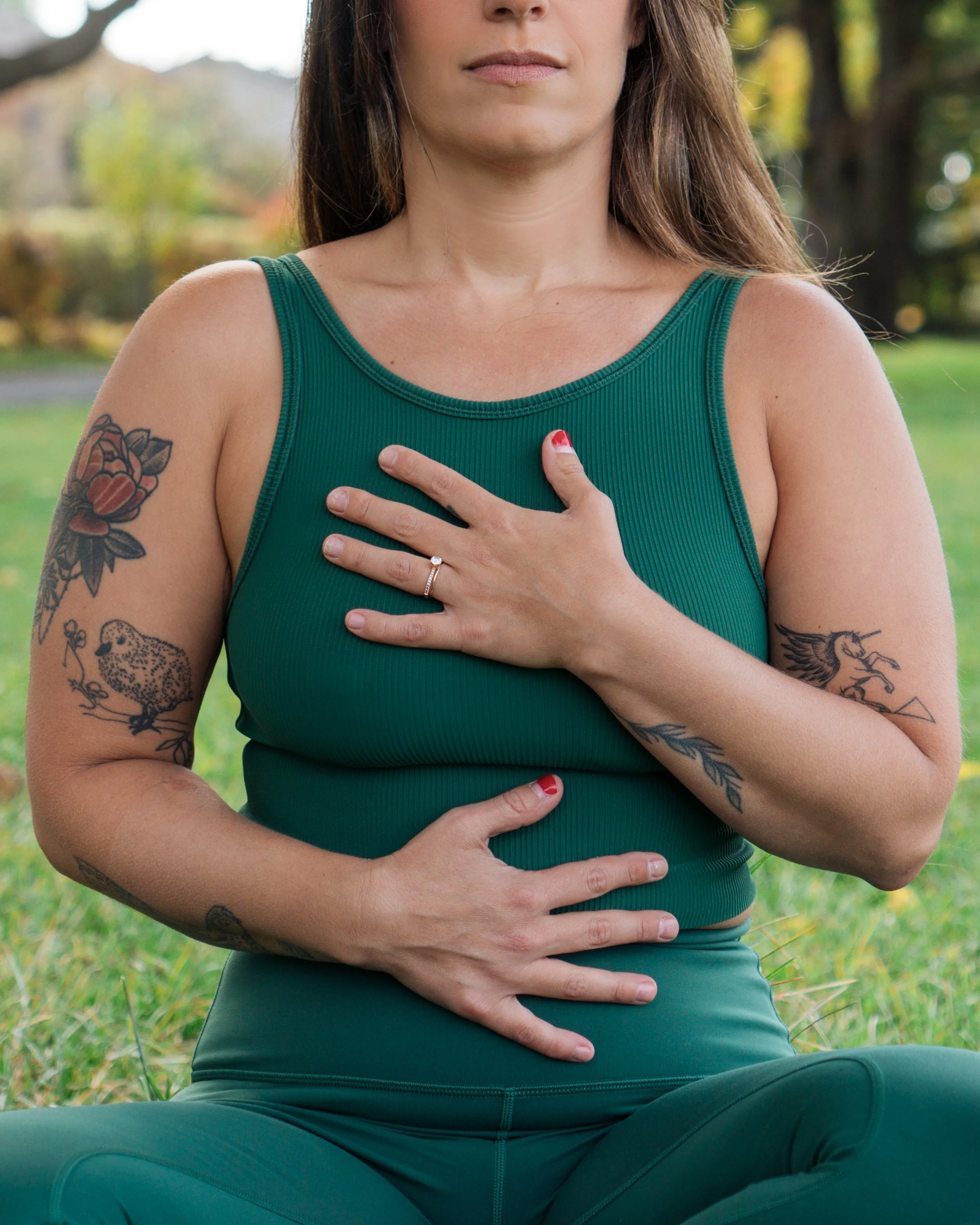 A woman practicing yoga or meditation outdoors, sitting cross-legged, with her eyes closed, wearing a sleeveless green top and matching leggings. She has tattoos on her left arm and her right hand placed over her heart.