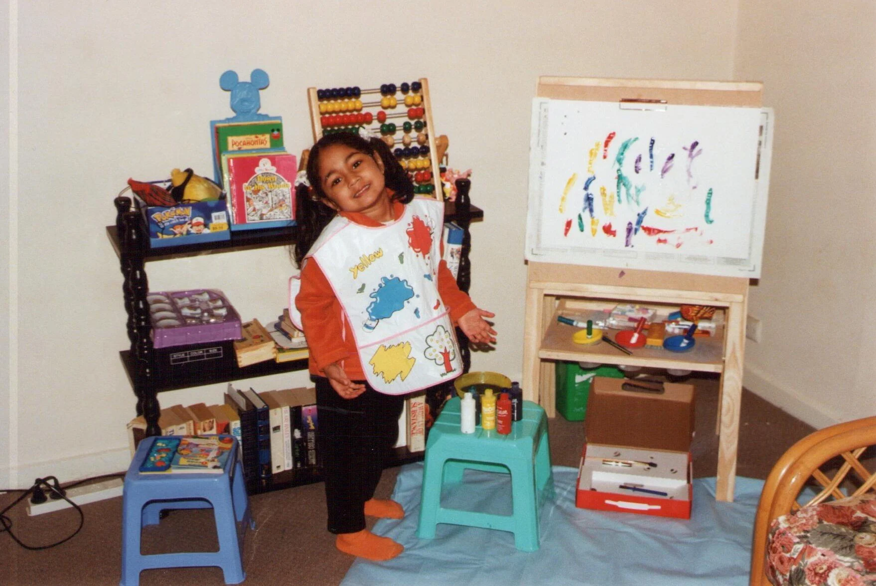 A young girl with dark hair, wearing an orange long sleeve shirt and artist apron, standing in front of an art station with colorful paints, brushes, and an abstract painting on a whiteboard. She is smiling and gesturing towards her artwork.