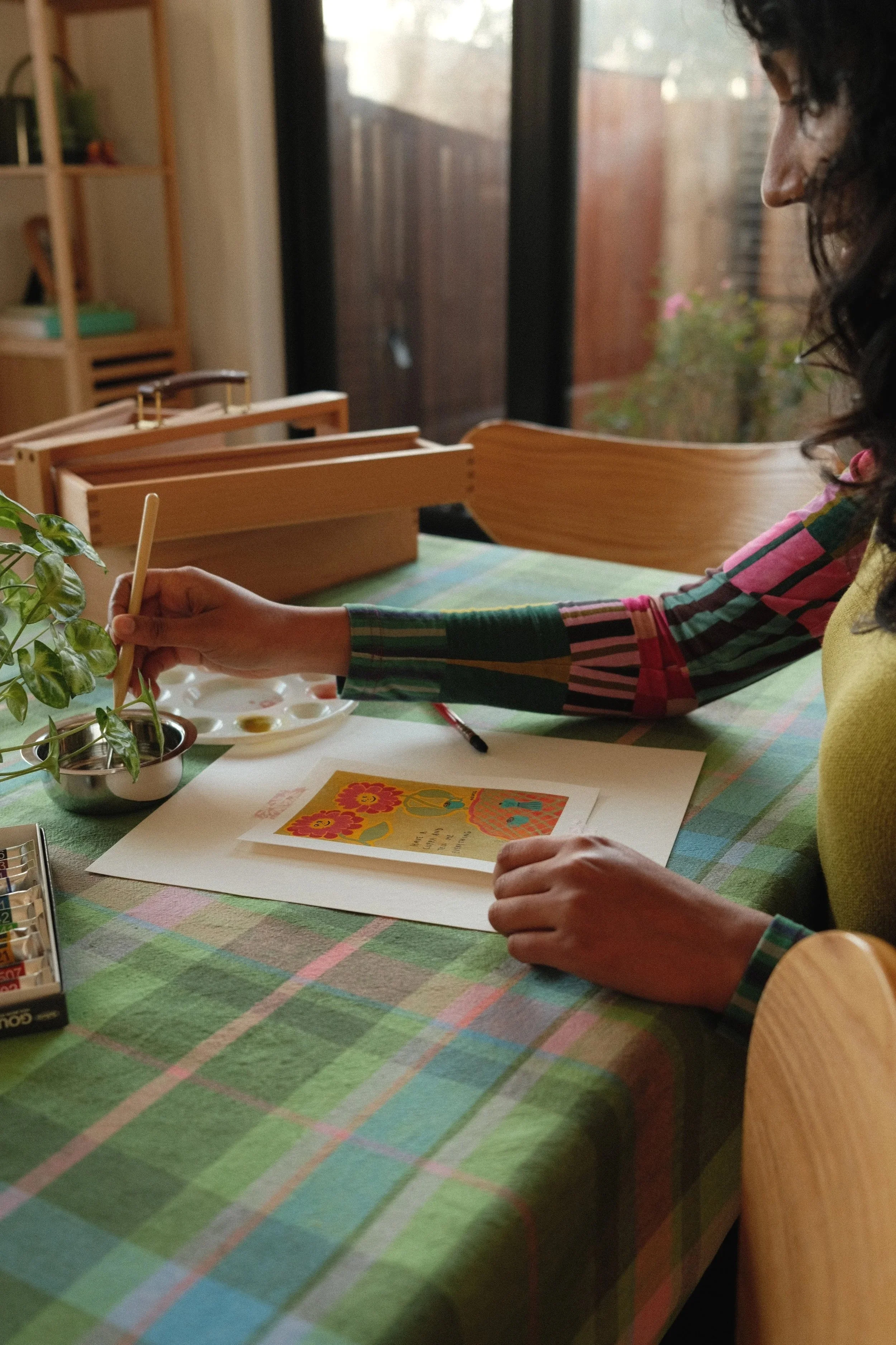 A person wearing a yellow shirt with a rainbow-and-stripe pattern on the sleeves is sitting at a table covered with a green checkered tablecloth. They are holding a paintbrush and working on a bright, colorful greeting card with flowers and cheerful designs. A potted plant is on the table. In the background, a wooden shelf holds books and decorative items, and sunlight filters through large window panes.
