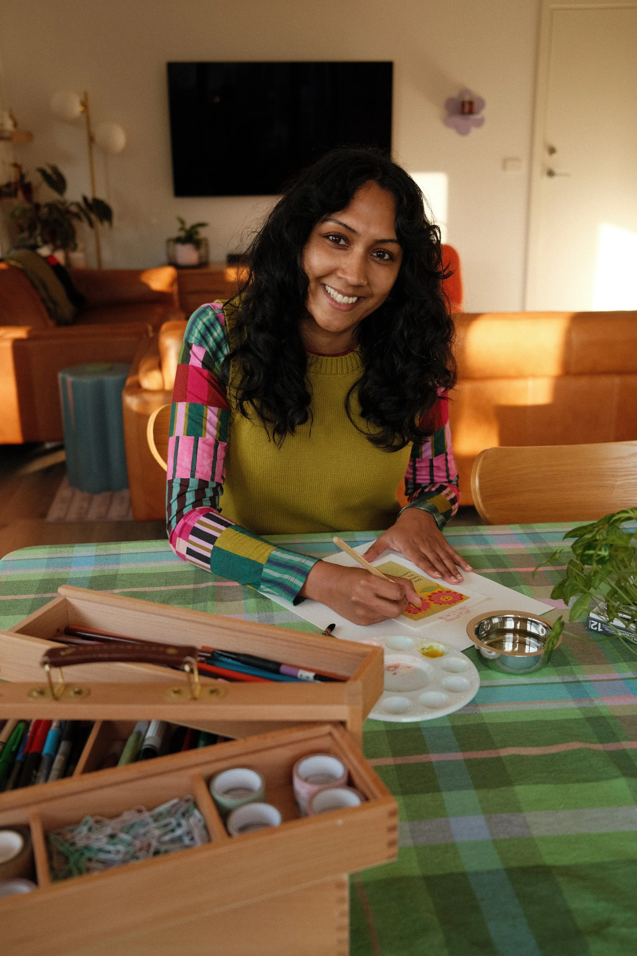 Smiling woman with dark curly hair sitting at a table with art supplies, including paint and markers, in a cozy living room.