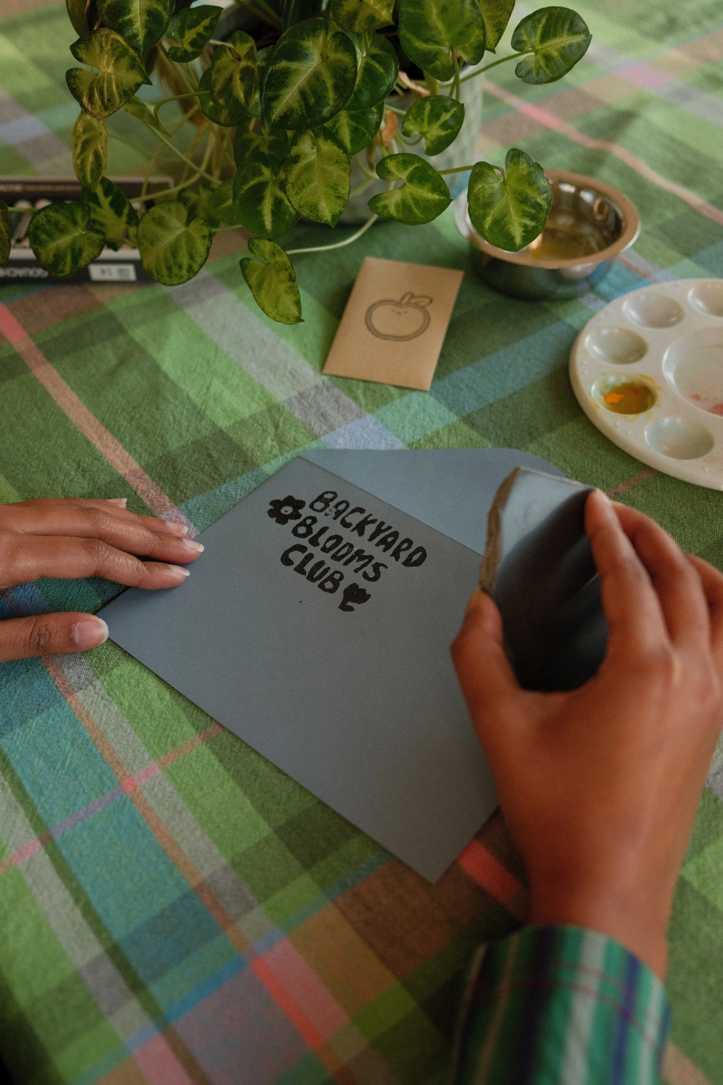 A blue envelope being stamped with 'Backyard Blooms Club', with a paint palette and stationary in the background.