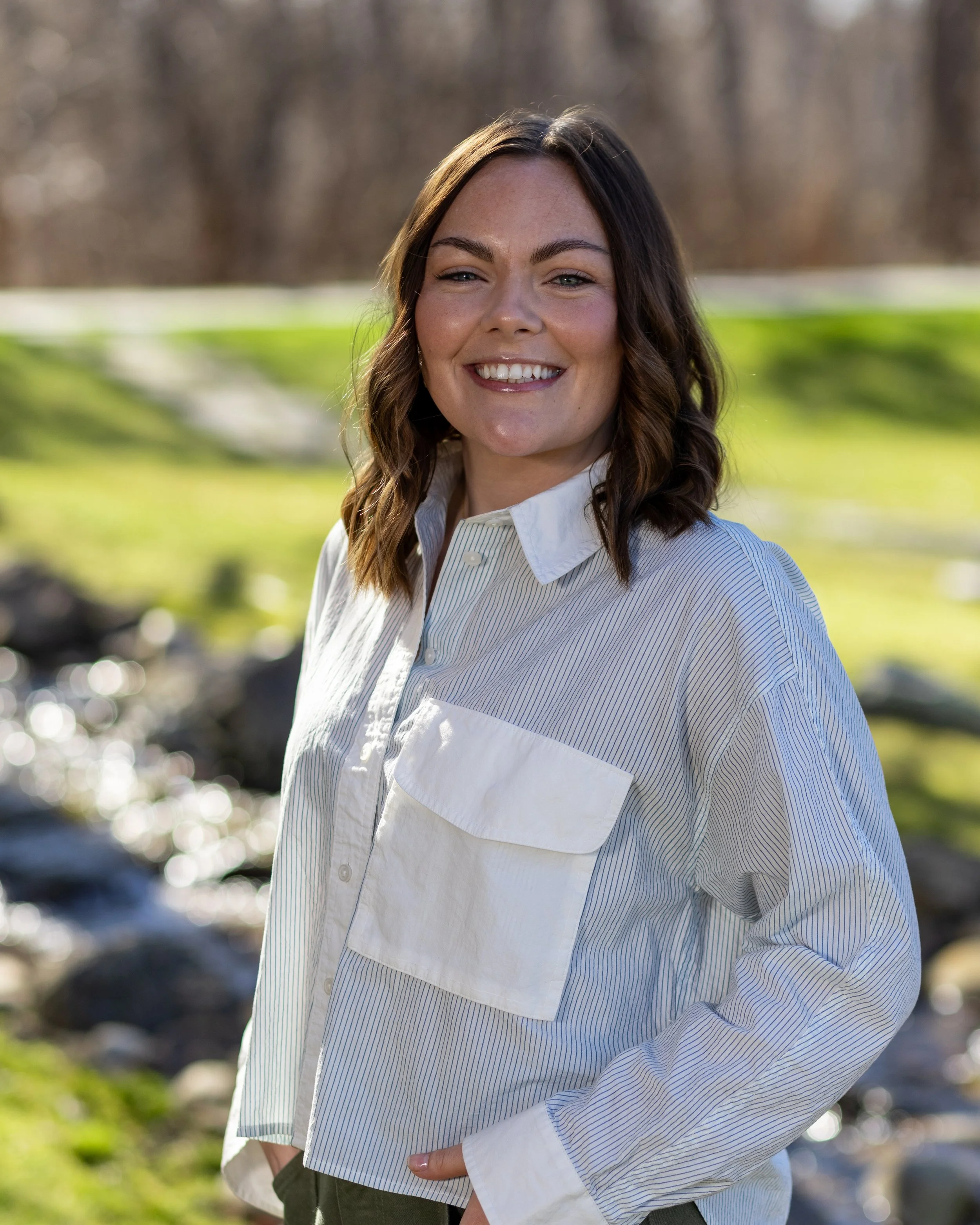 A smiling woman with long brown hair, wearing a white and blue striped shirt, standing outdoors with a blurry background of grass, trees, and a stream.