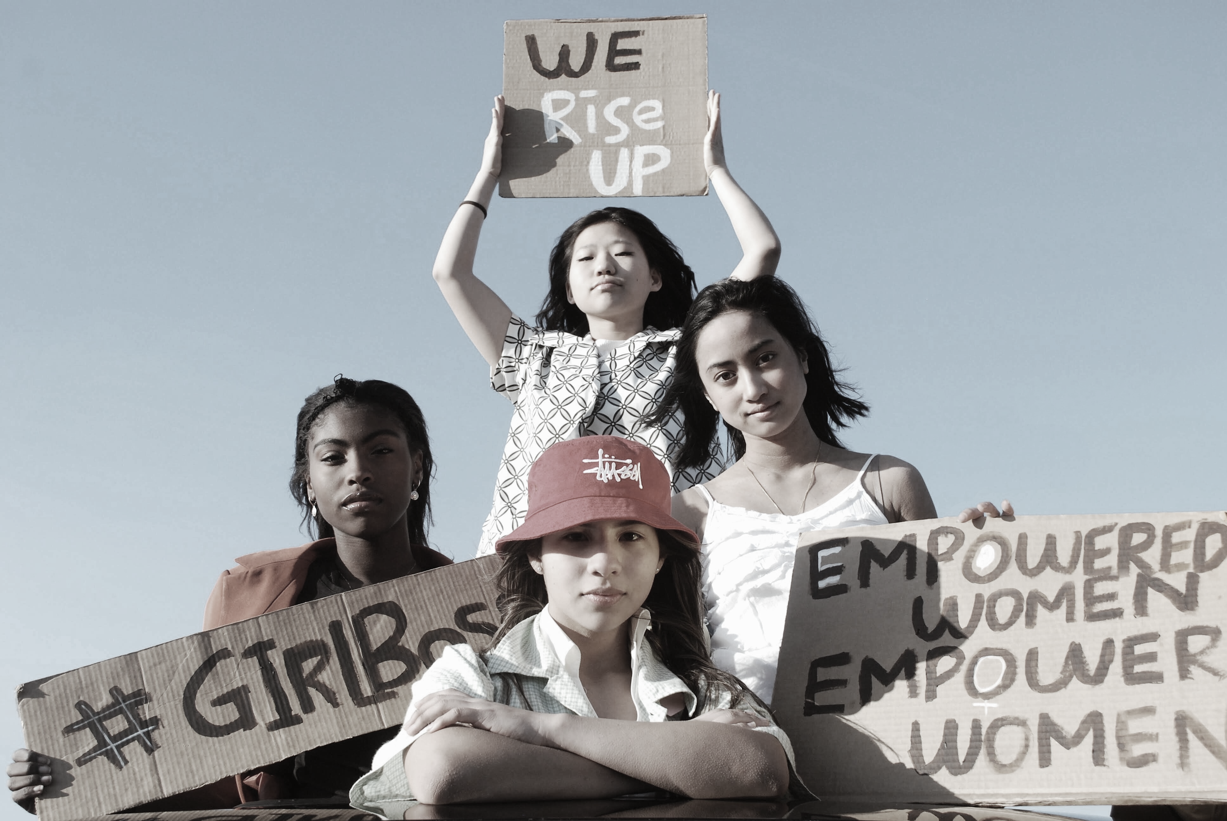 Group of diverse women and girls holding protest signs, one sign says 'WE RISE UP' and another hashtags '#GIRLS' and 'EMPOWER' are visible. The women are standing outdoors against a clear sky.