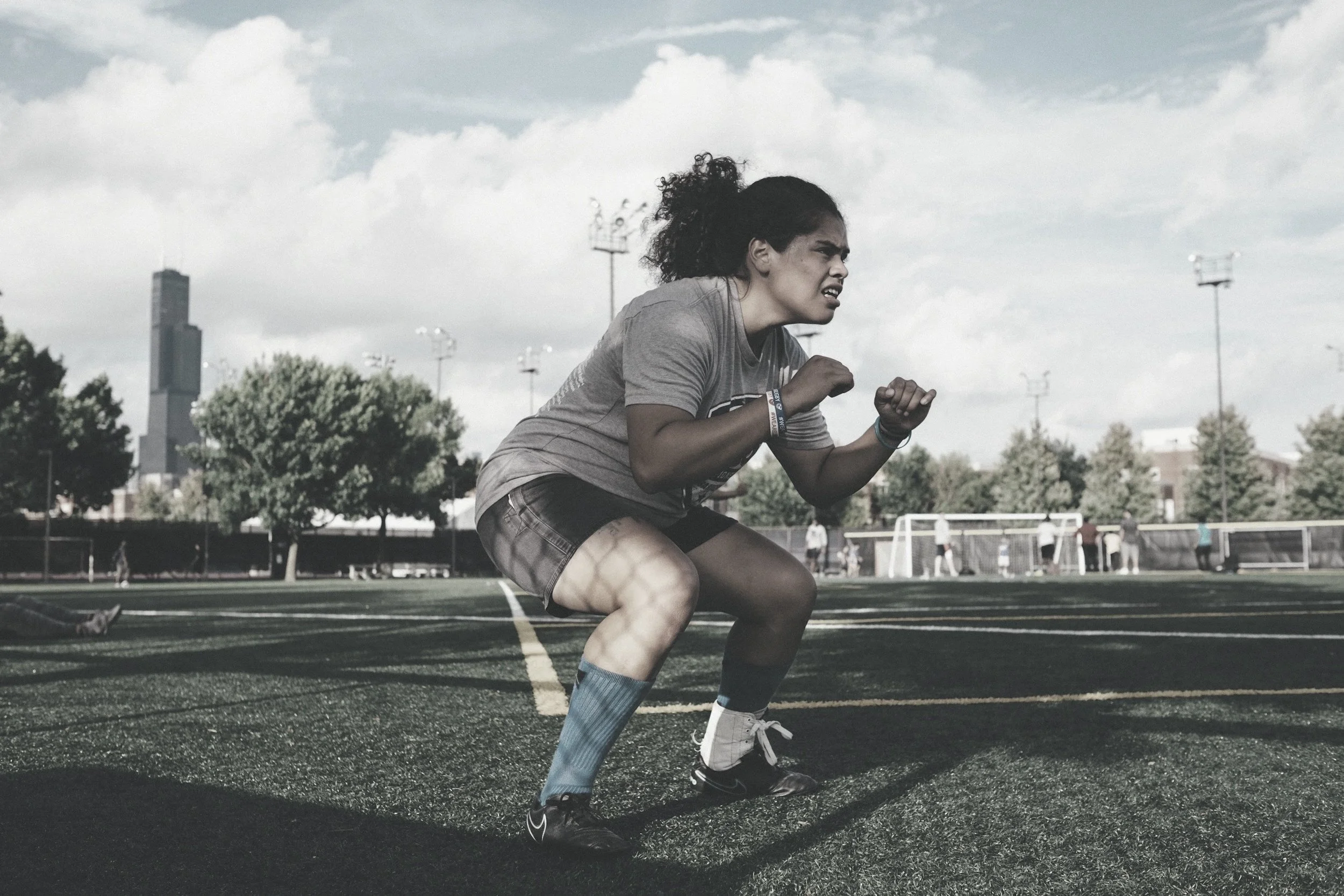 A young woman with curly hair wearing a gray t-shirt and shorts is crouched down on a running track, preparing to sprint. There are trees, athletes, and a city skyline in the background under a cloudy sky.
