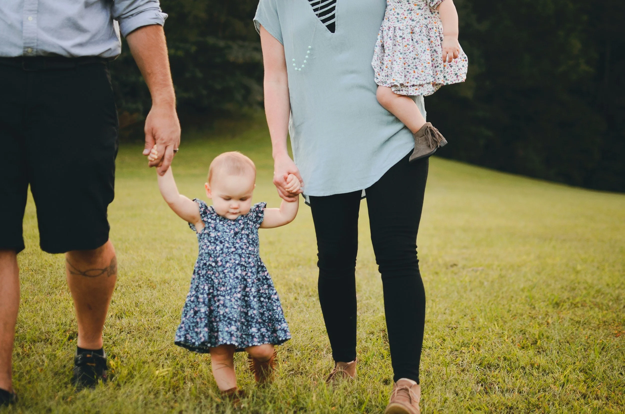 Young family with 12 month twins walking through grass. Mom has one baby on her hip, and she and dad are each holding a hand of the other twin as she toddles through the grass.