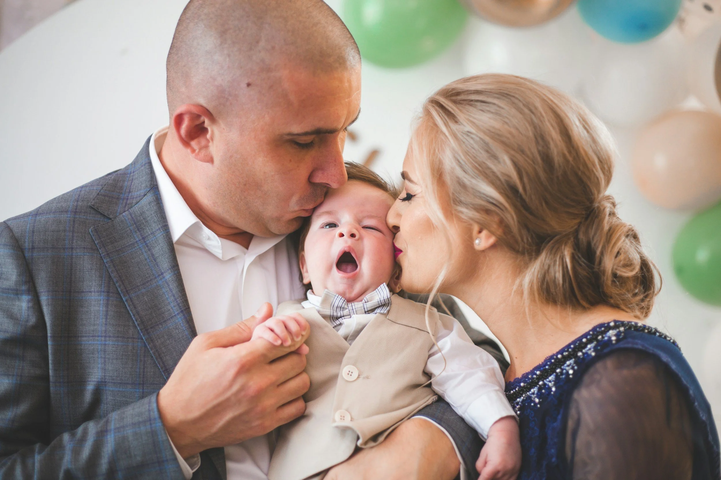mom and dad holding baby boy between them, each kissing him on the side of the head