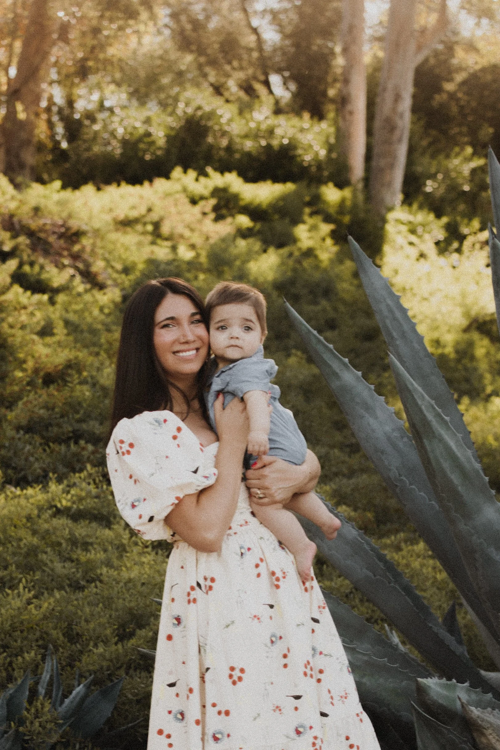 A woman with dark hair smiling and holding a young boy with short light brown hair outdoors near large agave plants, surrounded by green trees in warm sunlight.