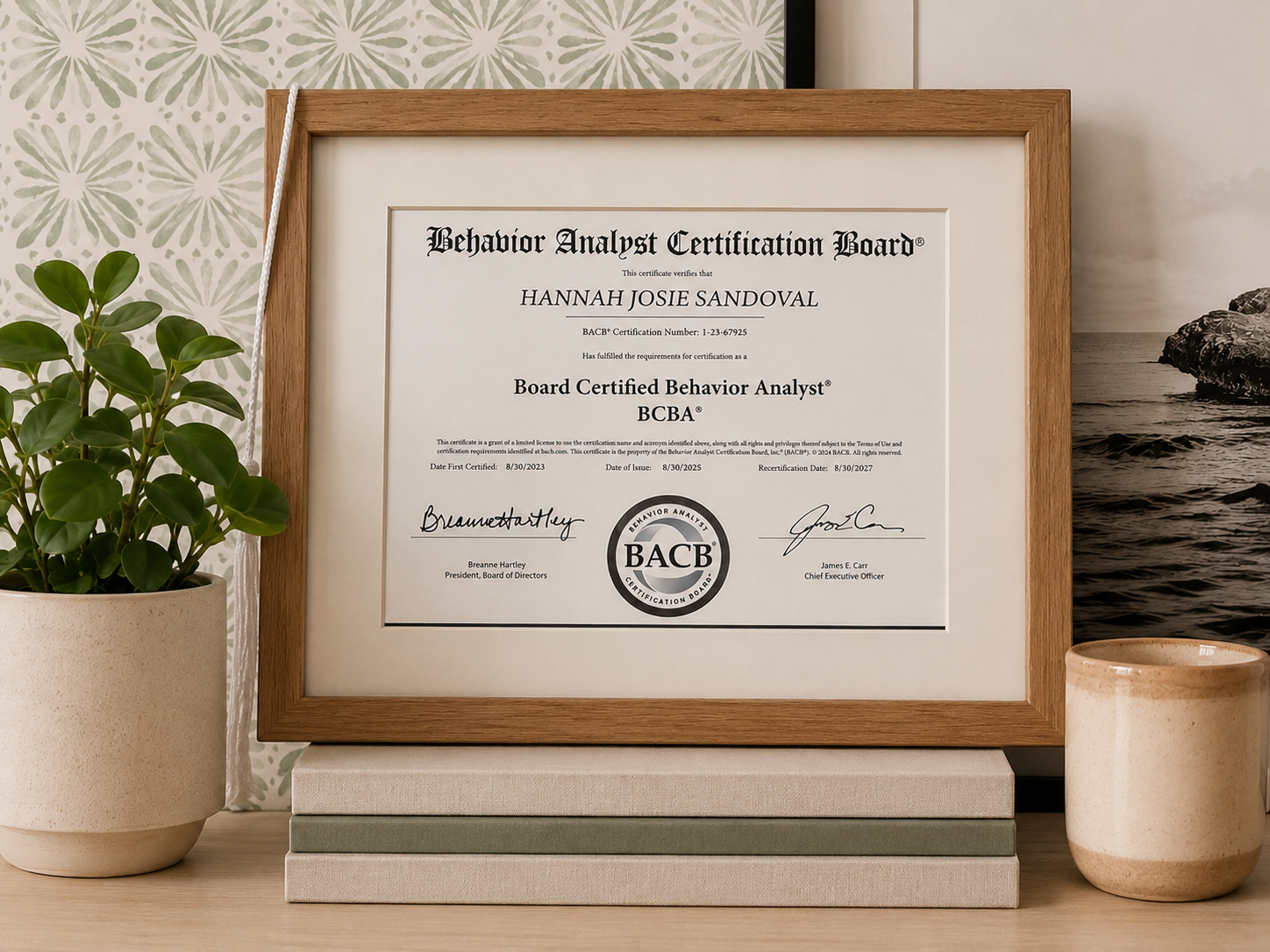 Certificate of achievement from the Behavior Analyst Certification Board awarded to Hannah Josie Sandoval for becoming a Board Certified Behavior Analyst, displayed in a wooden frame on a desk with books, next to a potted plant and a candle, with a decorative wallpaper and a photo of the ocean in the background.