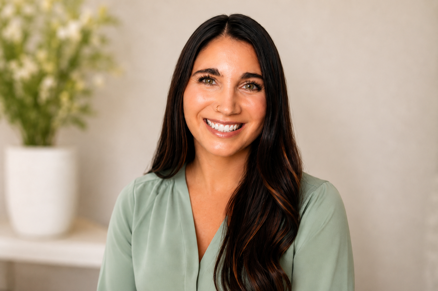 A smiling woman with long dark hair, wearing a light green blouse, sitting indoors with a blurred plant in the background.