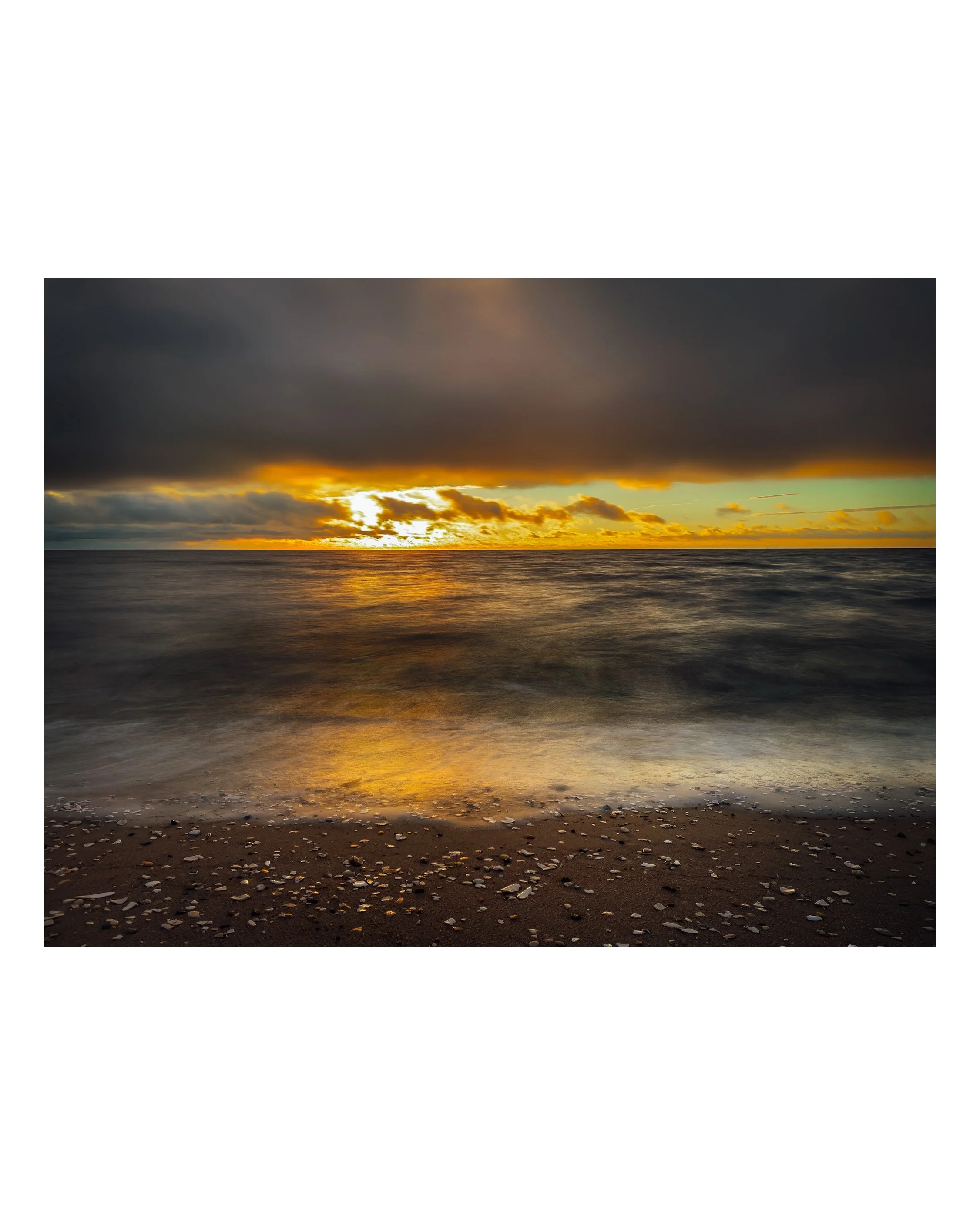 Sunset over the ocean with reflections on the water and scattered shells on the sandy beach in the foreground.