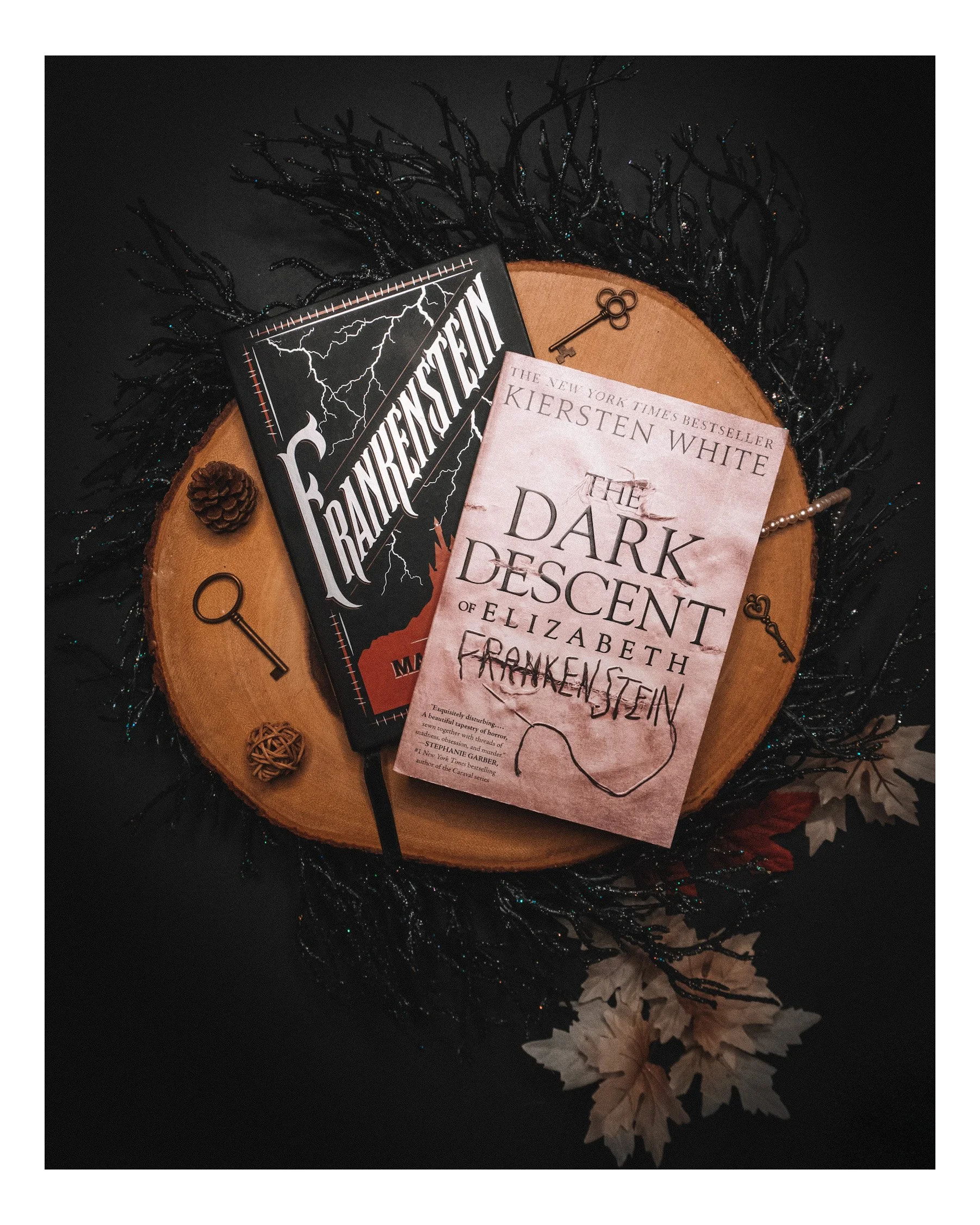 A wooden round platform with two books, a pinecone, a small pearl bracelet, and vintage keys, surrounded by decorations including tree branches, a pinecone, and faux leaves on black fabric with tiny glitter accents.
