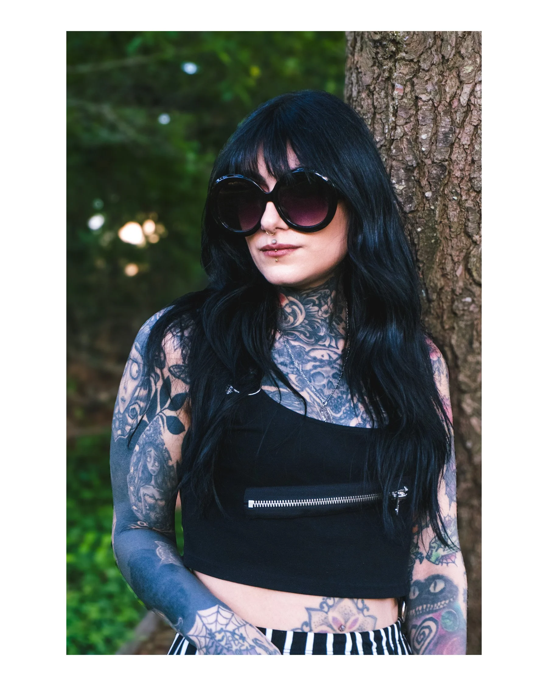 A woman with black hair wearing large sunglasses, a septum piercing, and tattoos on her arms and chest, standing outdoors with a tree trunk behind her and blurred greenery in the background.