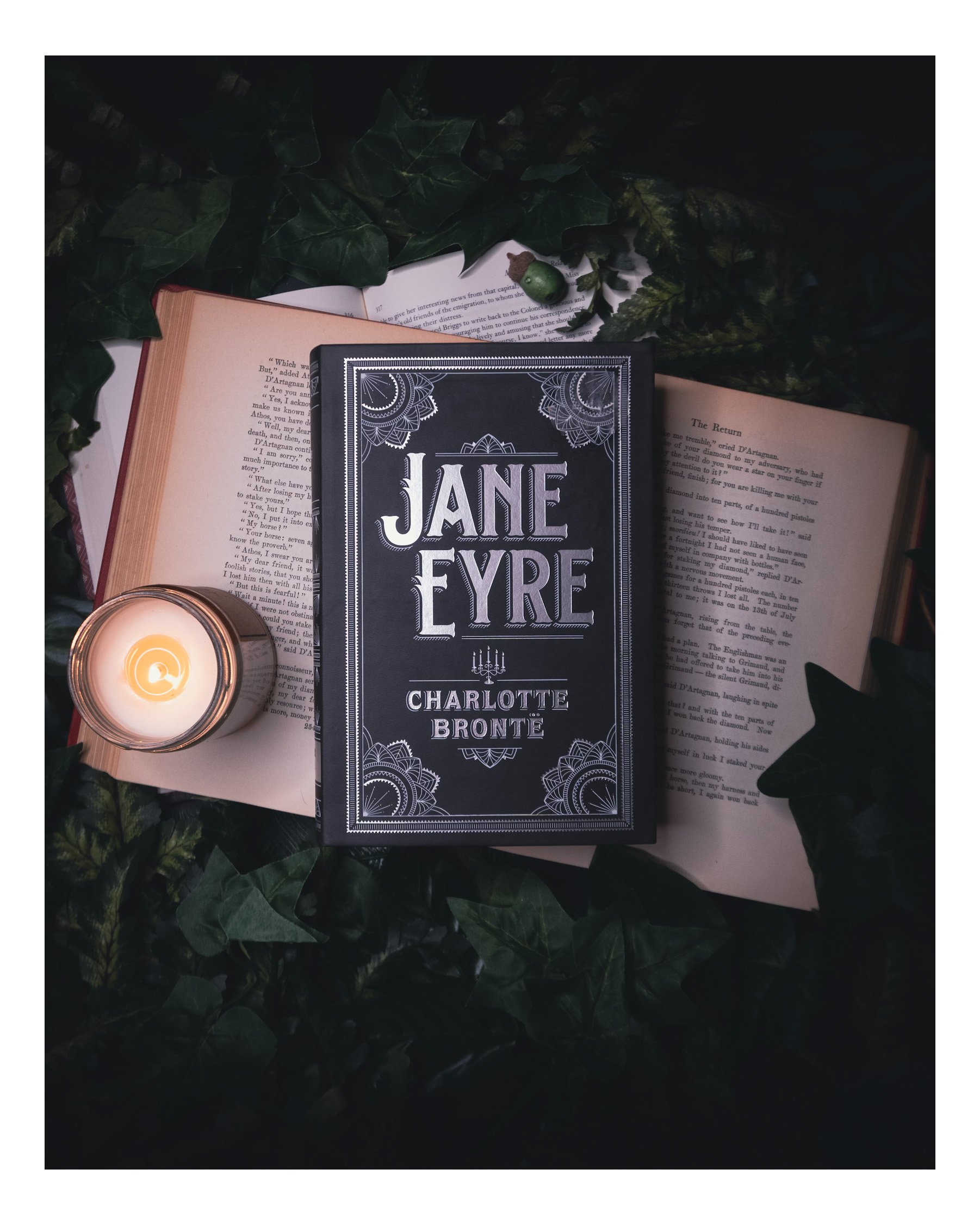 A book titled 'Jane Ever' by Charlotte Bronte is placed on top of other open books, surrounded by green leaves and a lit candle in a glass holder.