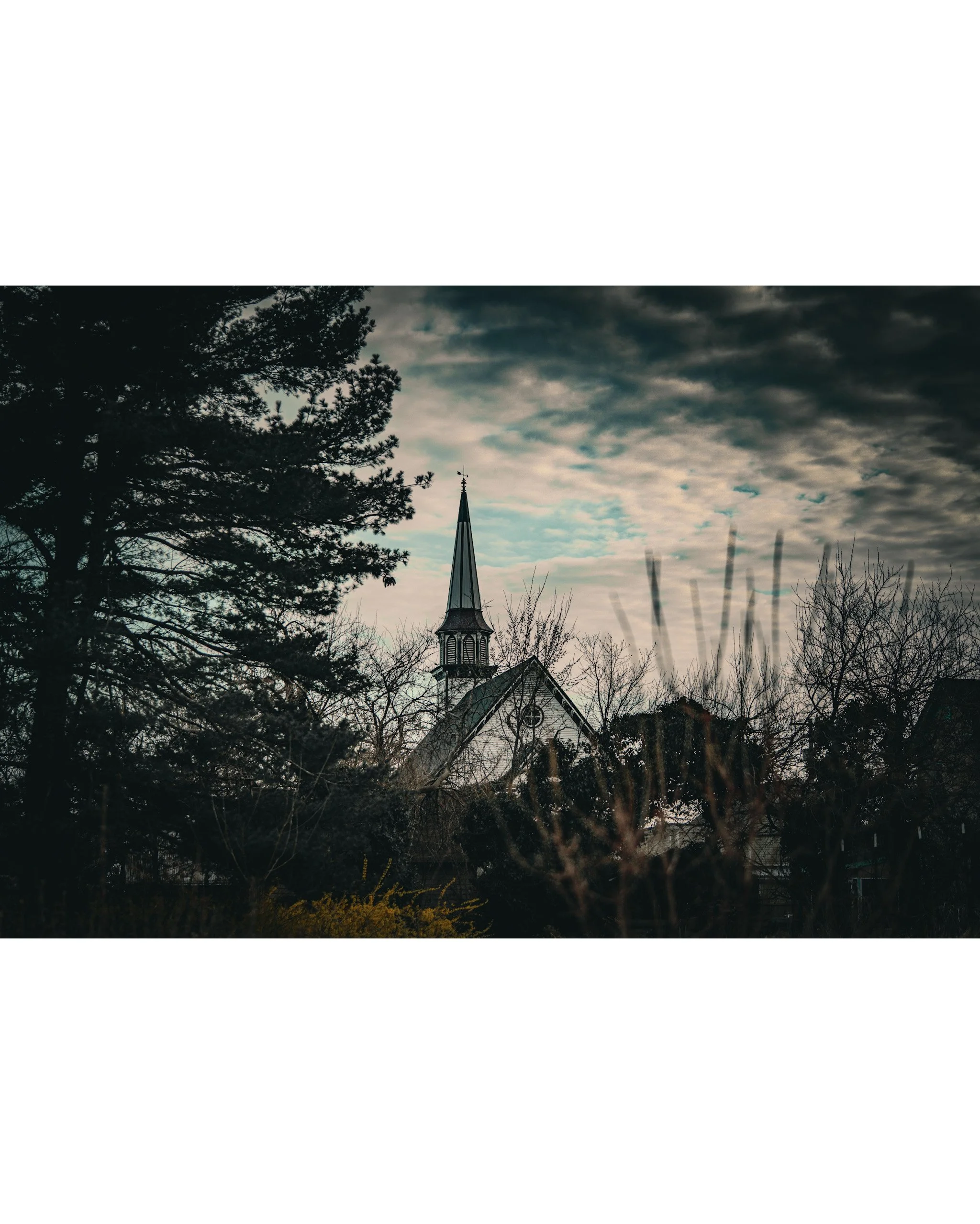 A church steeple surrounded by trees and bushes under a cloudy sky.