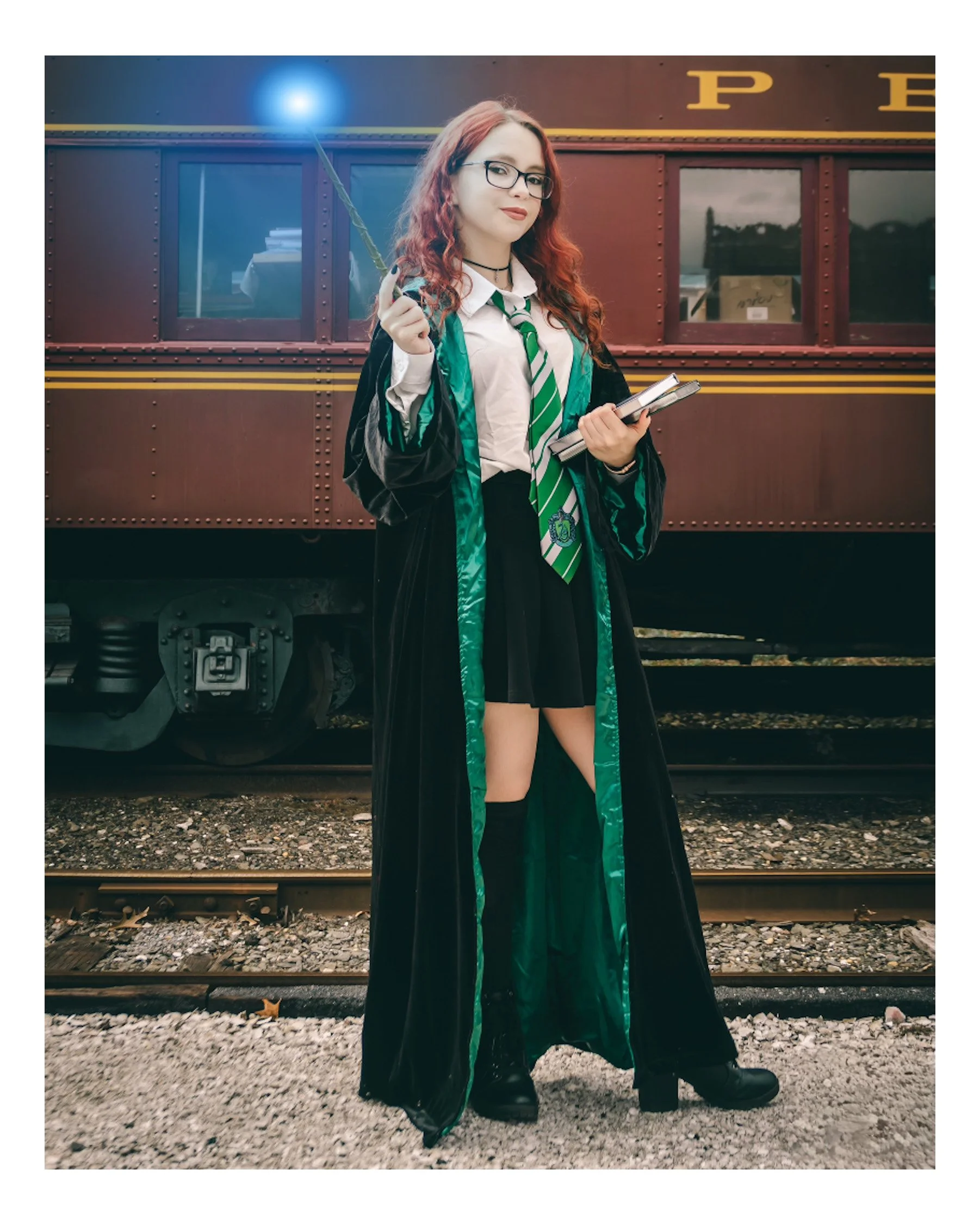 A young woman with red hair, glasses, and a black robe with green lining and Hufflepuff tie, holding a wand and a notebook, standing on train tracks in front of a maroon train car.