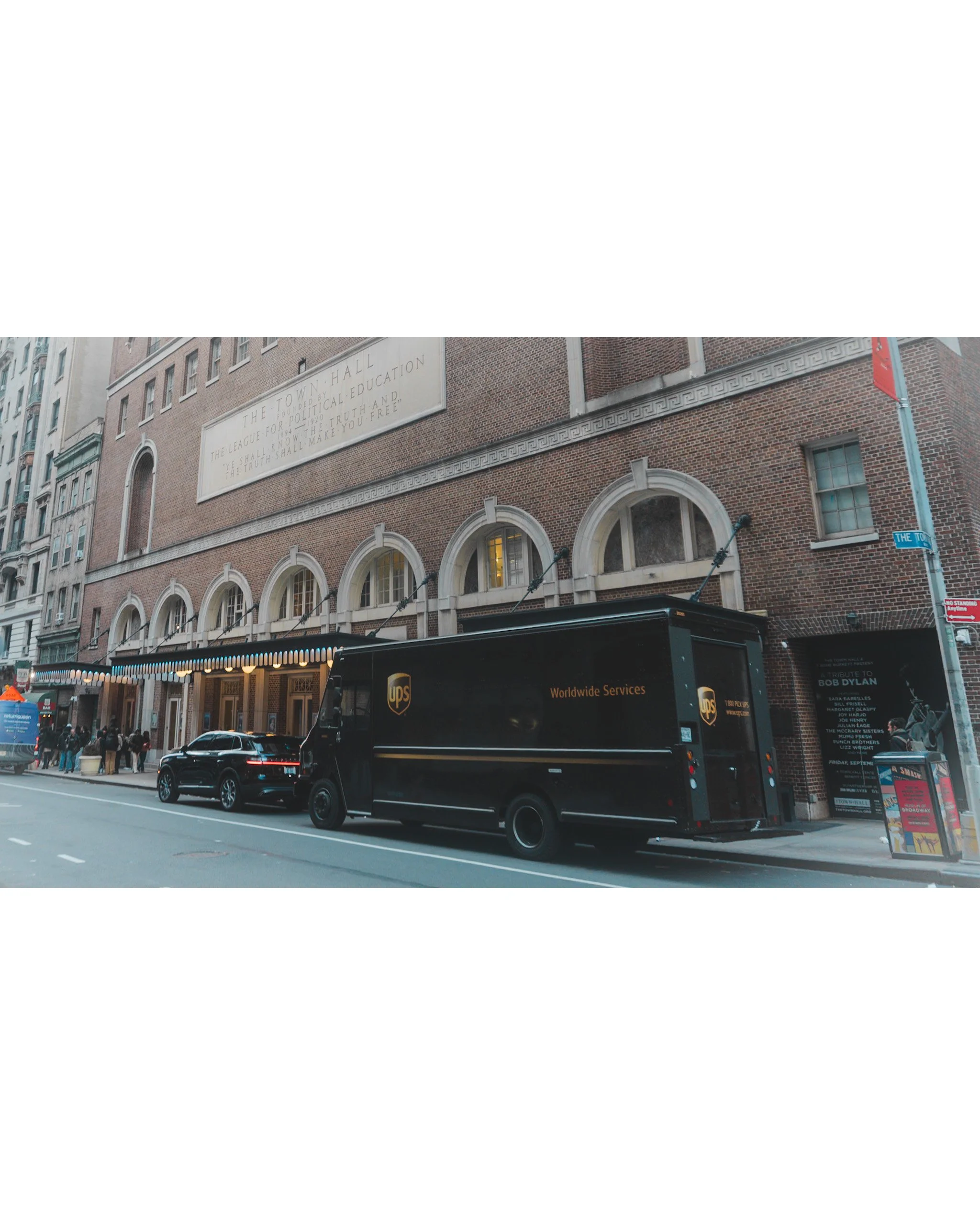 A city street scene with a large brick building featuring arched windows and a sign that reads 'The Town Hall.' There is a UPS truck with 'Worldwide Services' parked along the curb, with pedestrians walking on the sidewalk. There are various street s