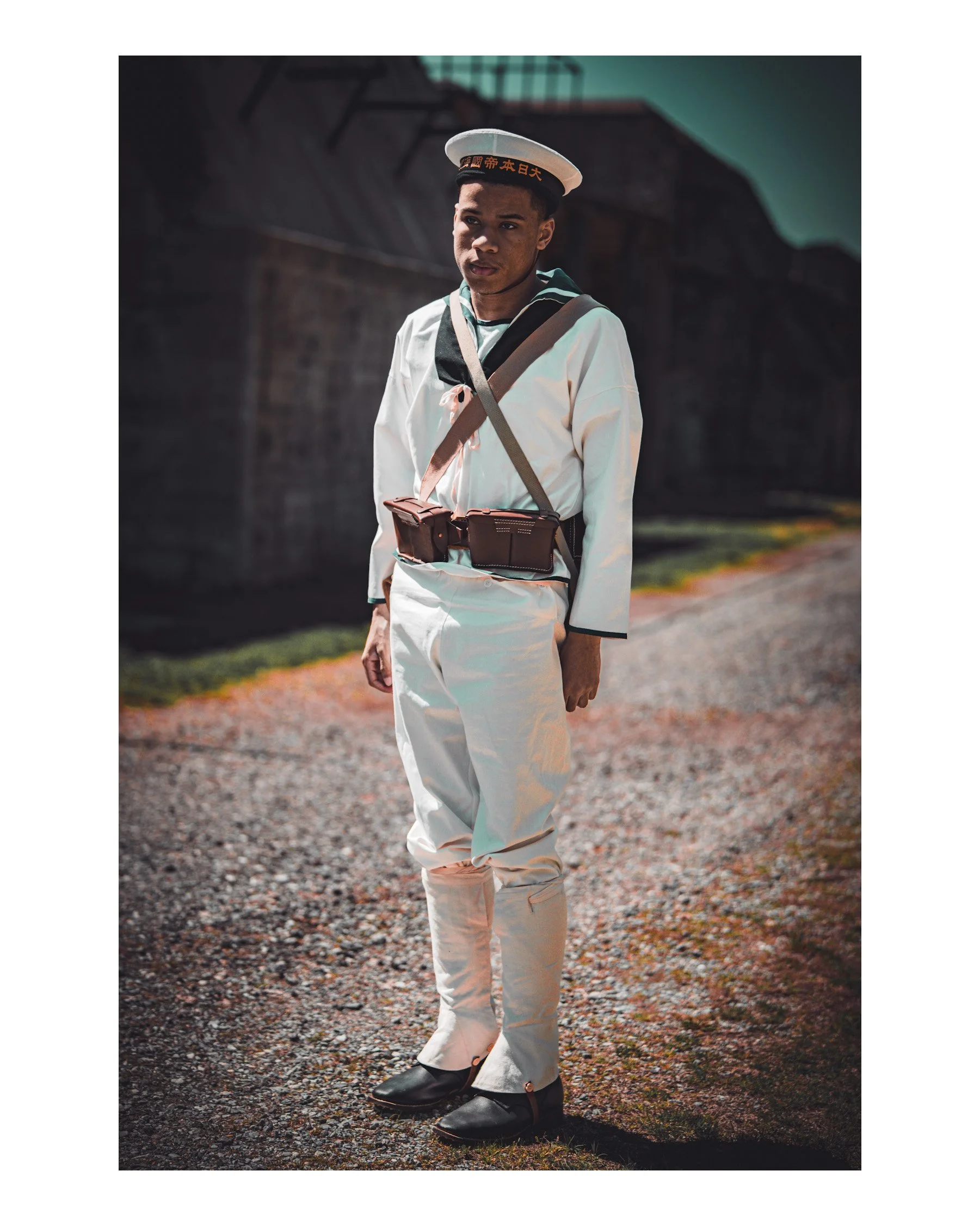 A young man in a sailor outfit standing outdoors on a gravel path with a dark building in the background. He is wearing a white sailor hat with Japanese characters, a white shirt with a blue collar, and beige pants. He has a shoulder bag and a pouch 