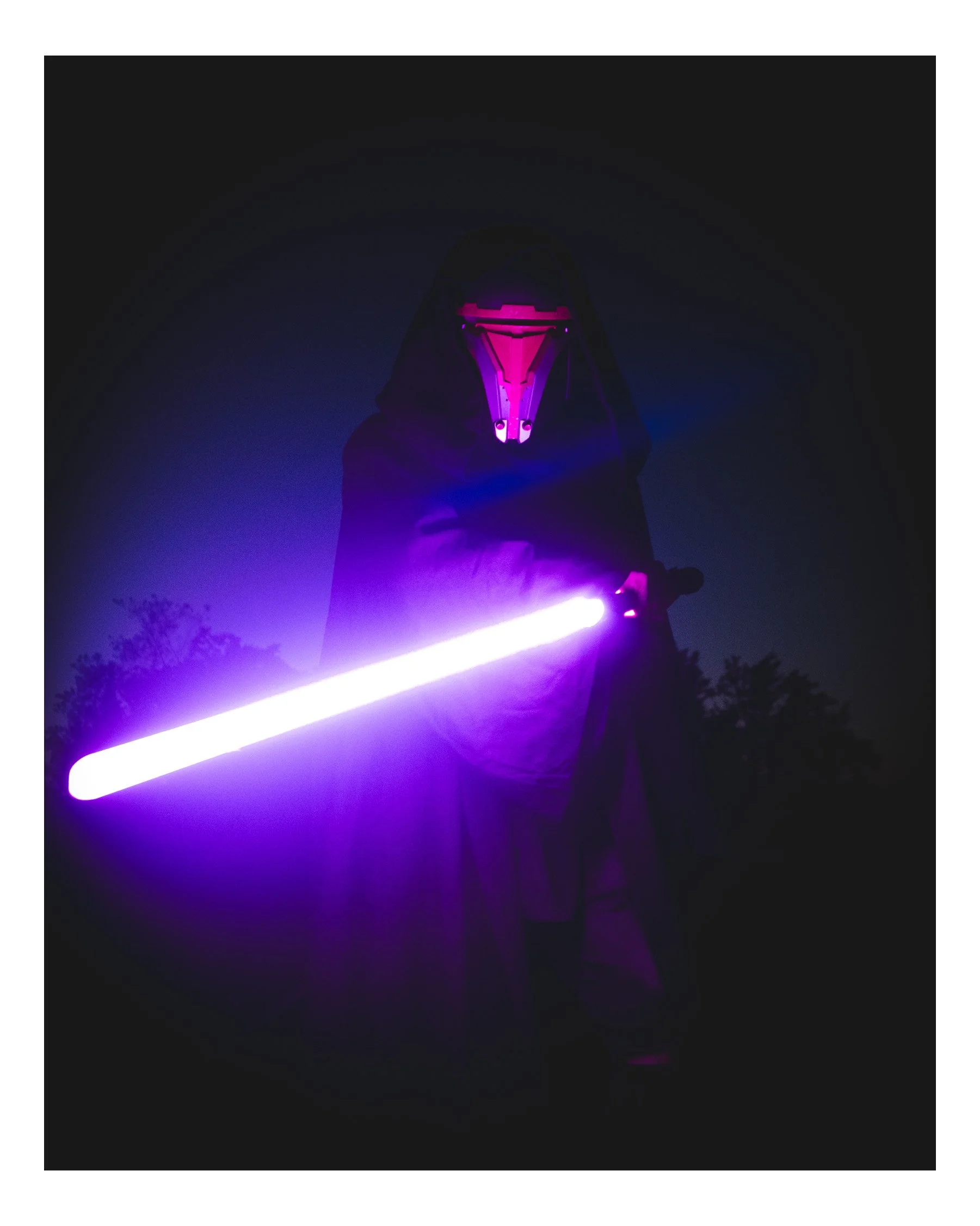 Person dressed as a Sith Lord from Star Wars, wearing a black hooded cloak and a shiny, intimidating mask, holding a glowing purple lightsaber in a dark environment.