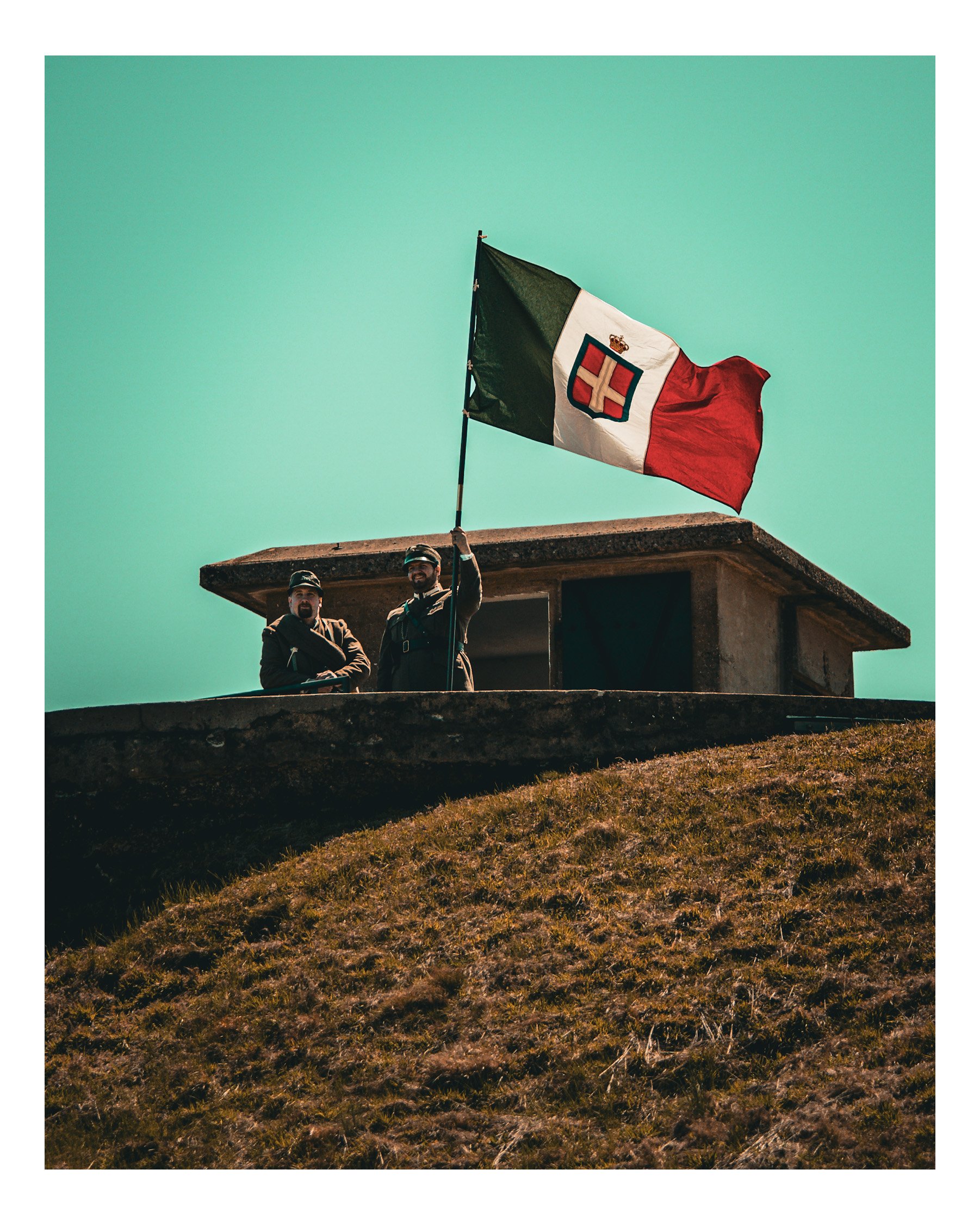 Two soldiers standing on a rooftop holding a large Italian flag against a clear sky with a grassy hill in the foreground.