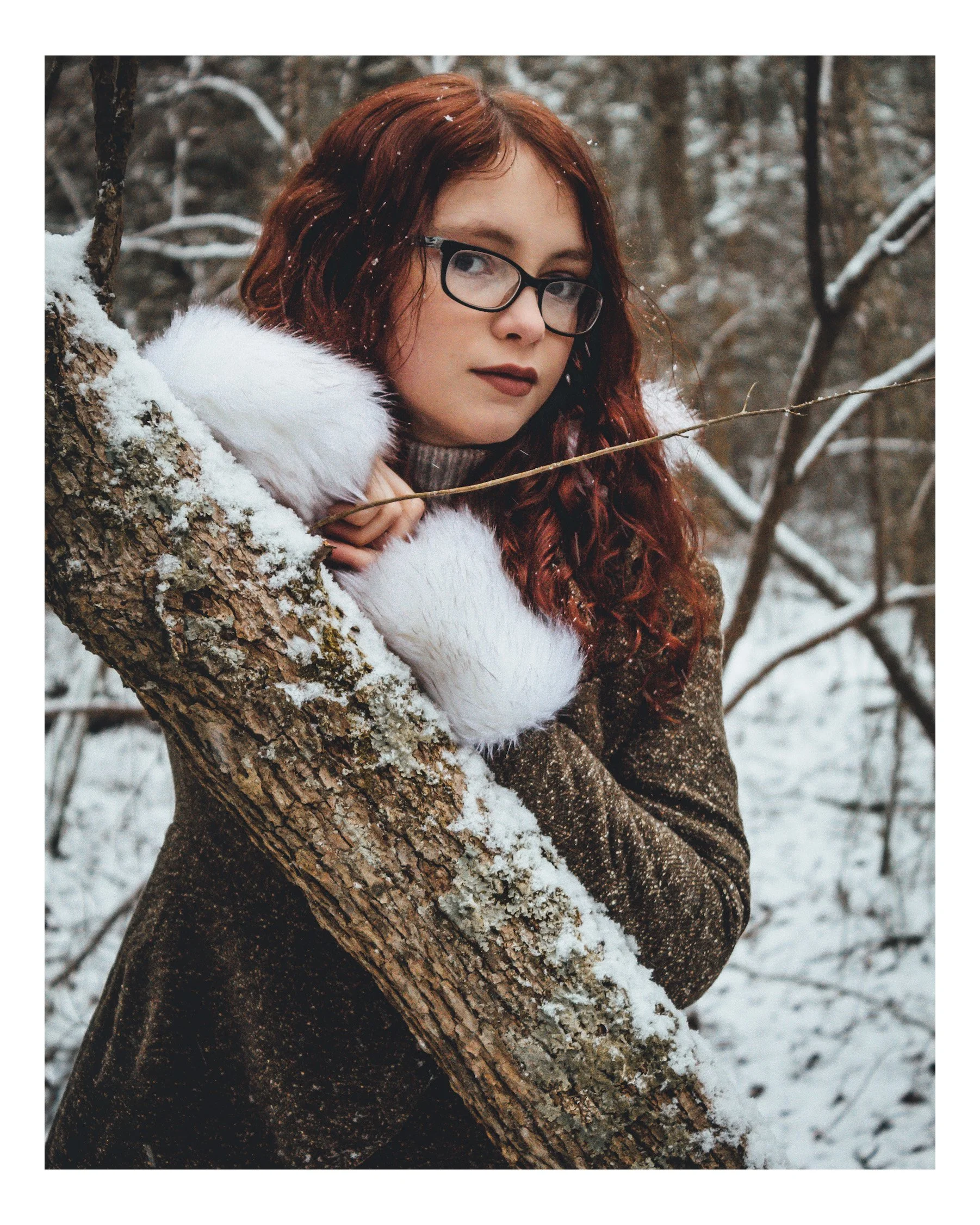 A young woman with red curly hair and glasses leaning against a snow-covered tree in a winter forest, wearing a brown coat with a white fur collar.