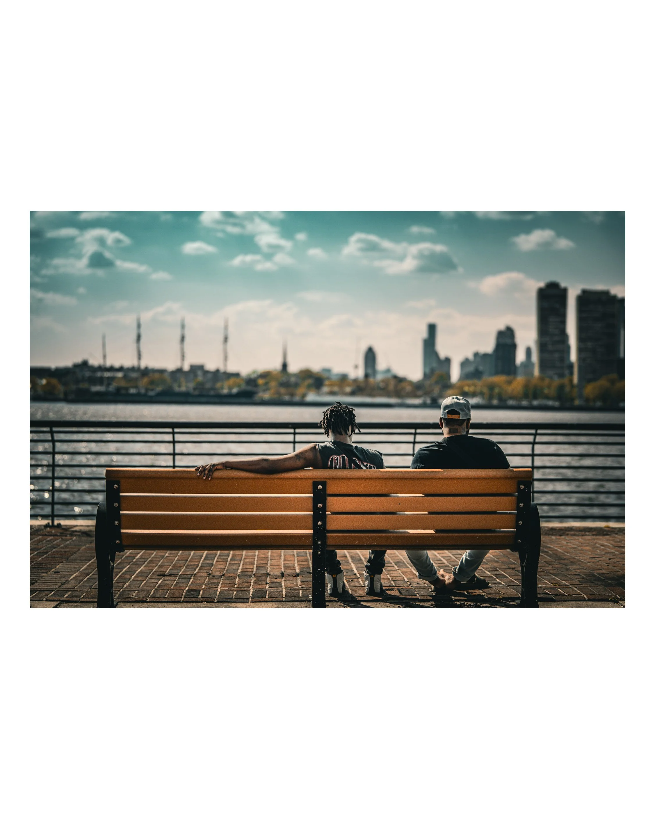Two people are sitting on a park bench by a river, with the city skyline in the background and a partly cloudy sky overhead.