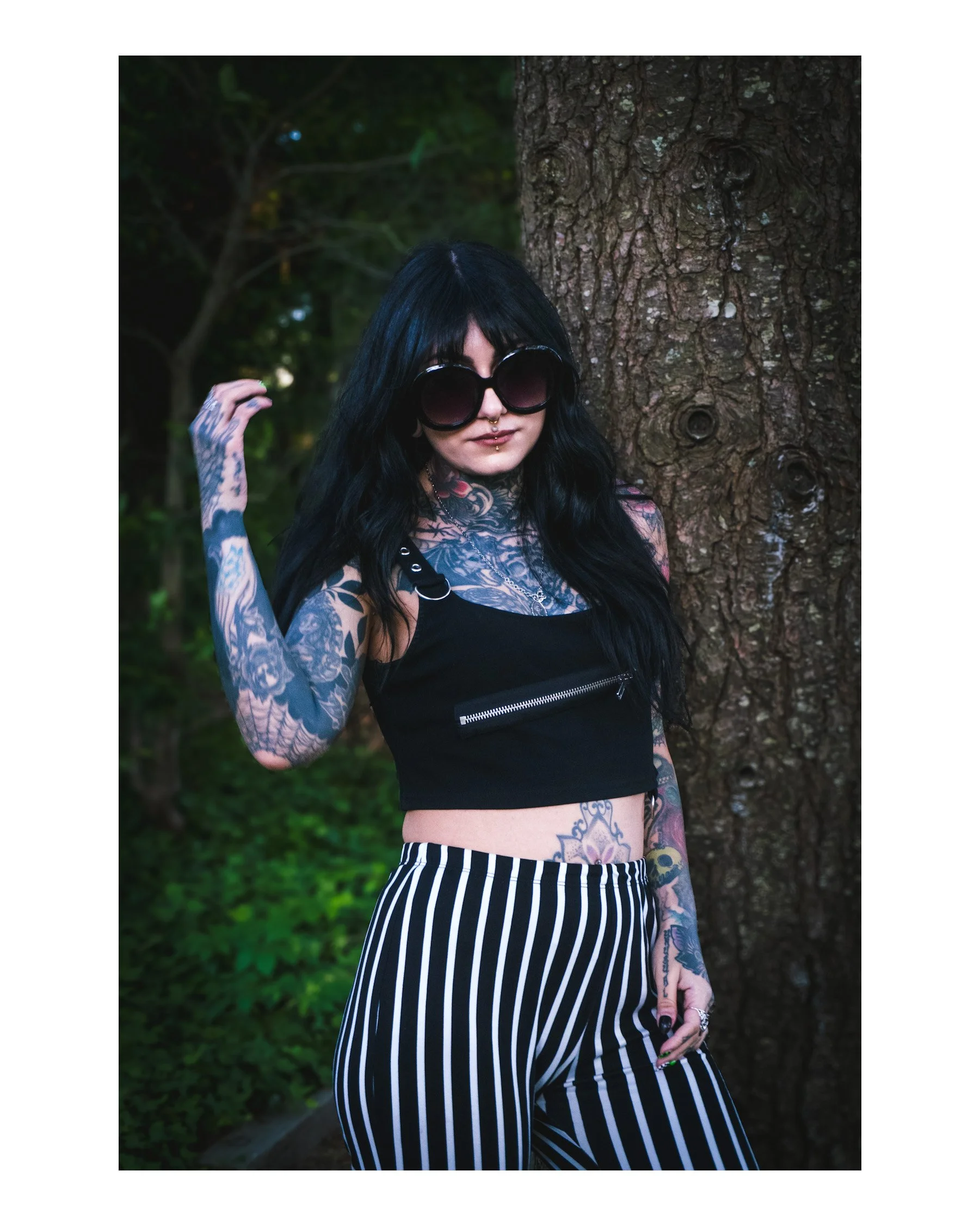 A woman with black hair, large dark sunglasses, and visible tattoos on her arms and torso stands outdoors in front of a tree with textured bark, wearing a black crop top and striped pants.