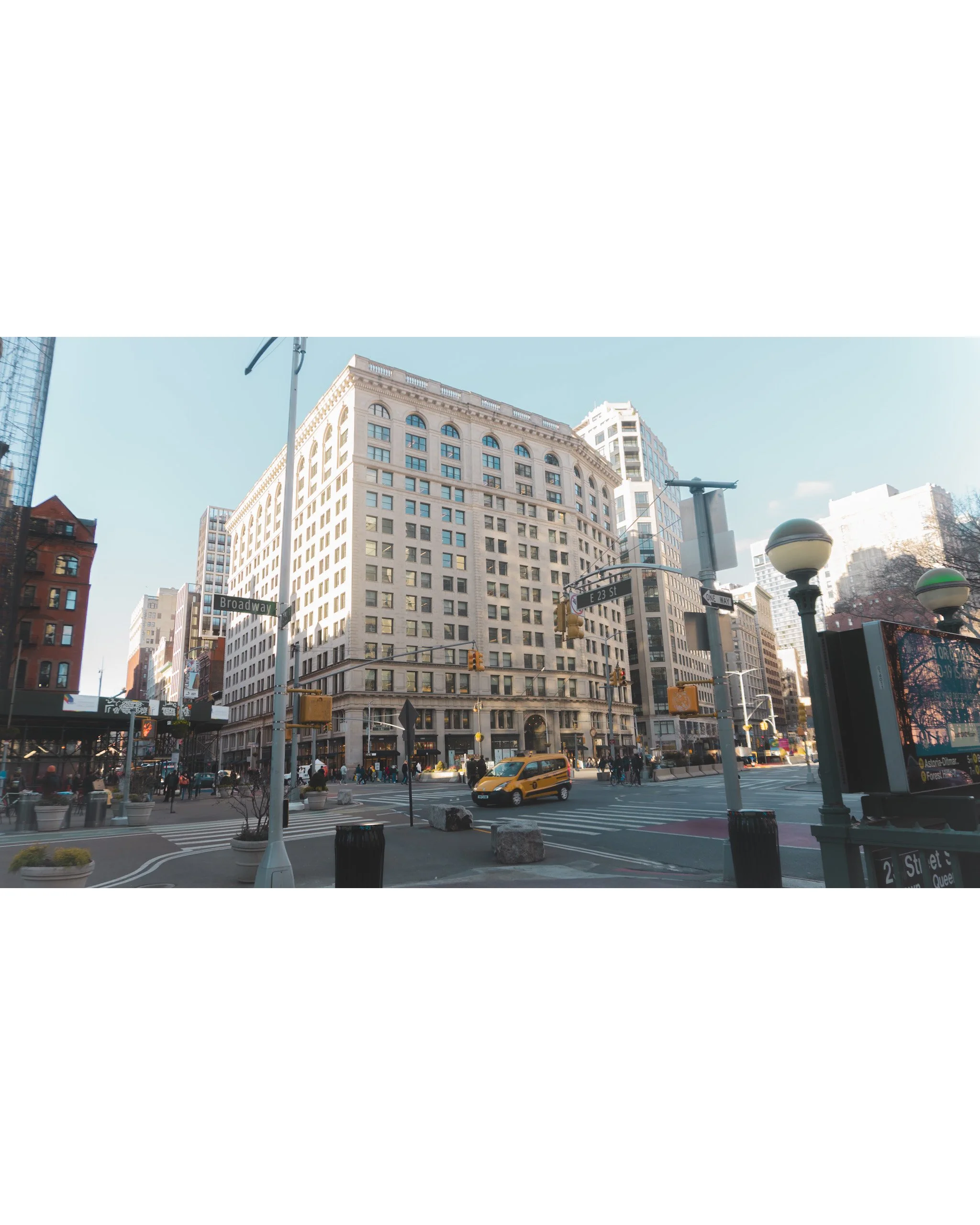 A busy city street scene in New York City with tall buildings, pedestrians, a yellow taxi, traffic lights, street signs, and street lamps.