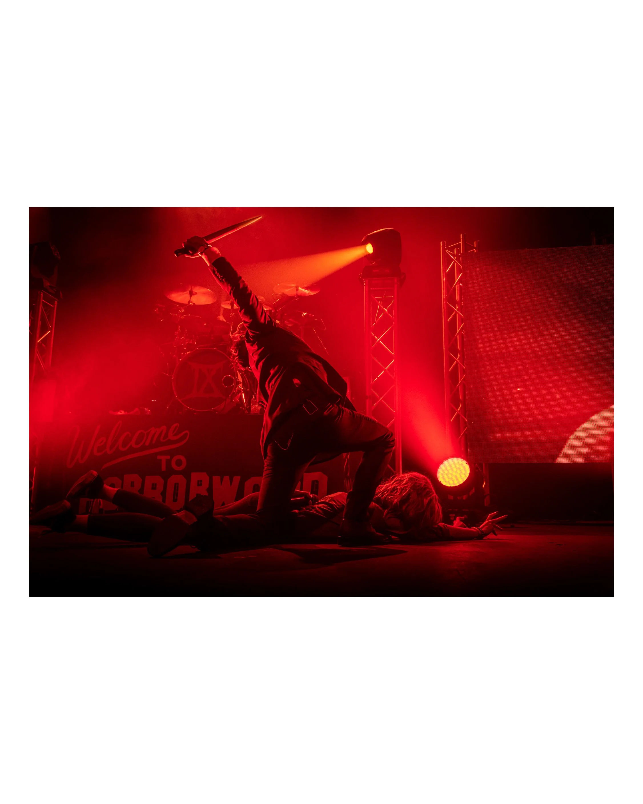 Performer on stage performing a death drop dance move, with one hand on the floor and the other holding a prop sword, illuminated by red stage lighting.