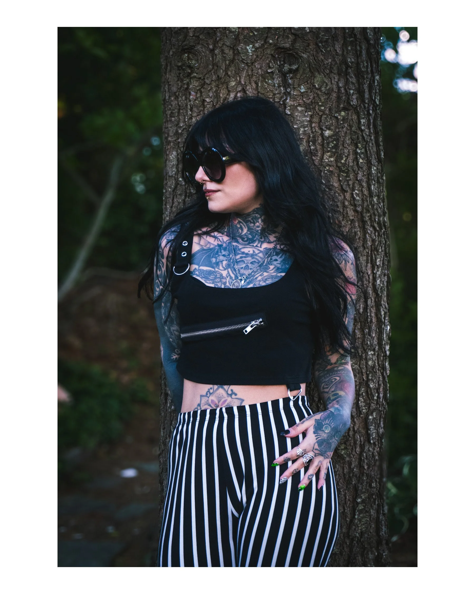 A woman with long black hair, dark sunglasses, and tattoos on her chest and arms leans against a tree in a wooded area, wearing a black striped skirt and a black top with metallic rings.