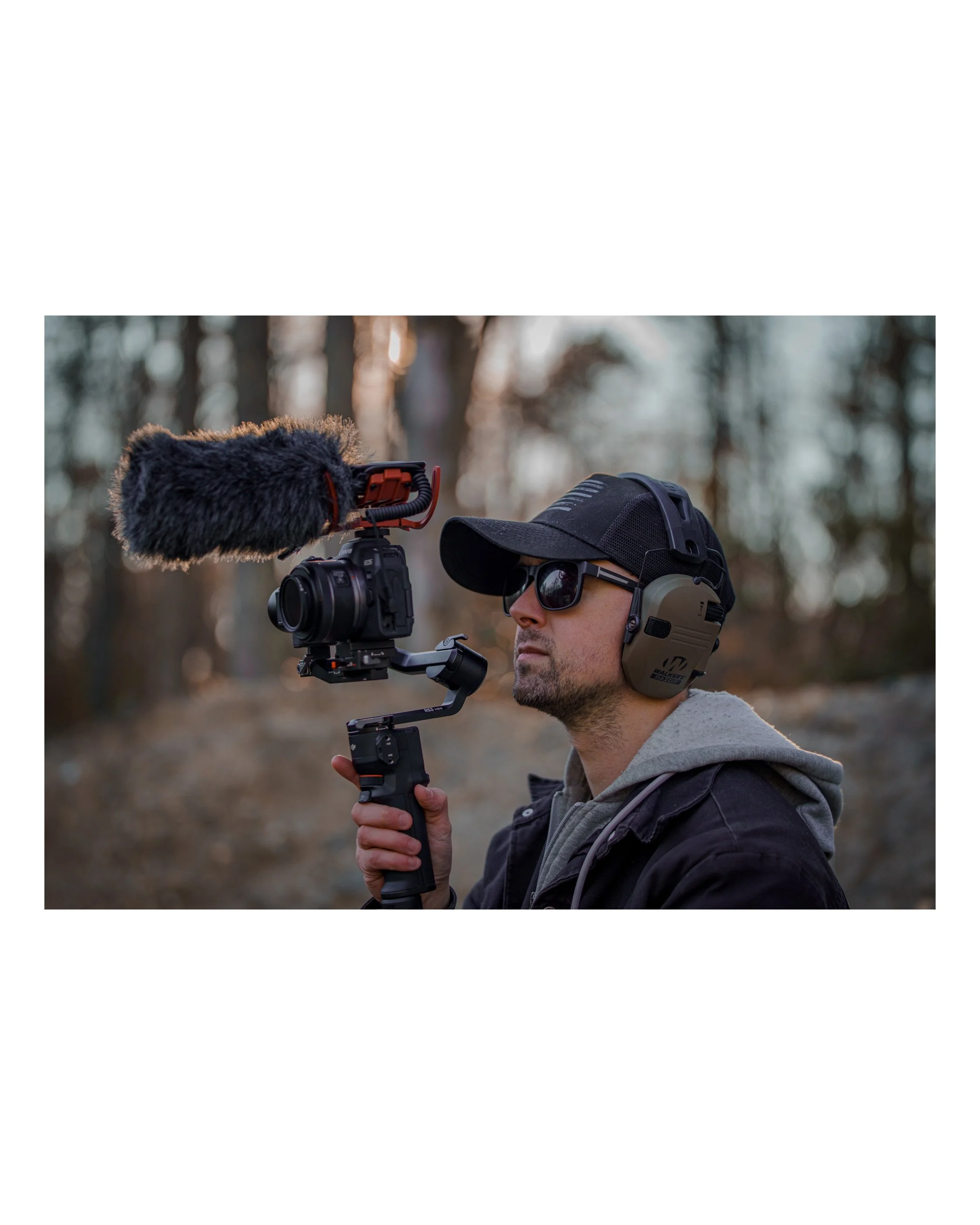 A man wearing sunglasses, a hat, and headphones, holding a camera stabilizer with a mounted microphone, outdoors in a wooded area with blurred trees in the background.