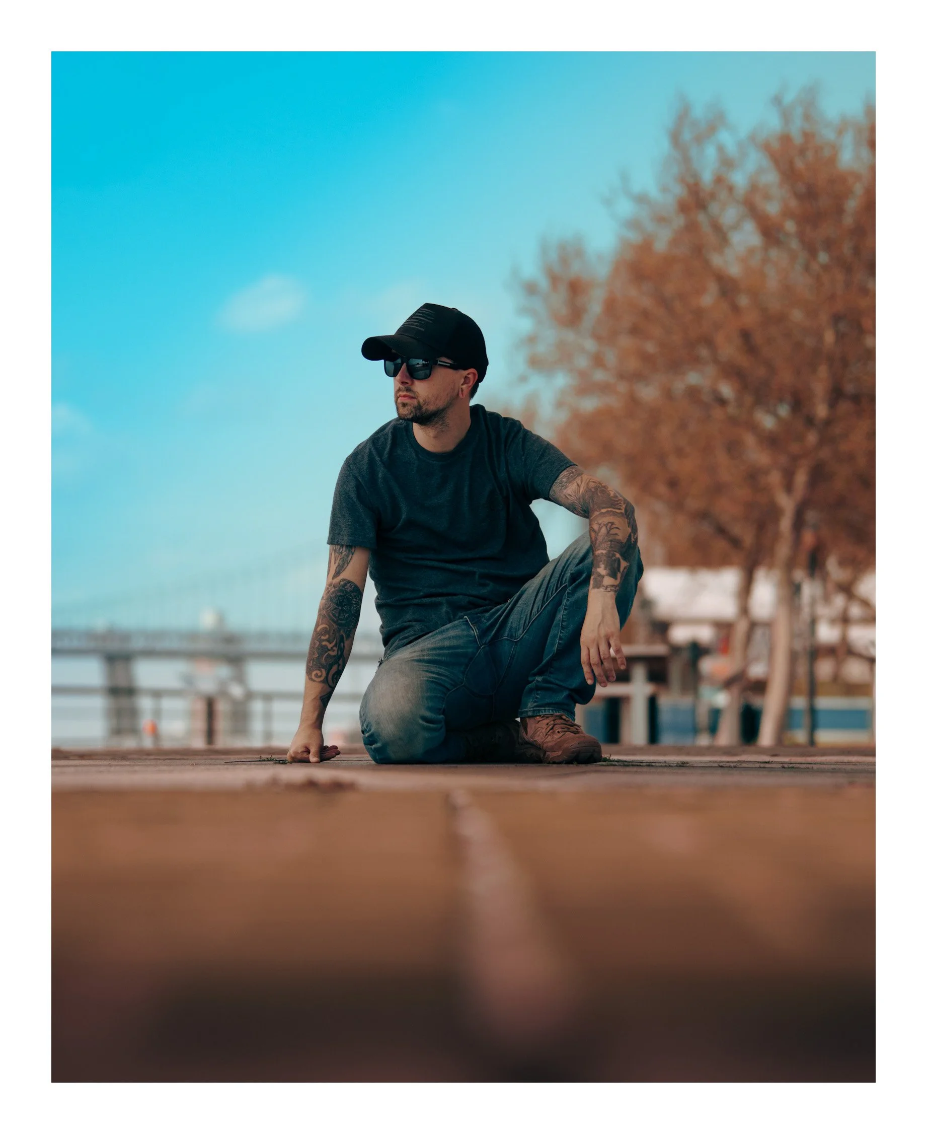 A man in sunglasses, a cap, and a dark t-shirt with tattoos on his arms kneeling on a wooden pier outdoors with a blurred background of trees and sunset sky.