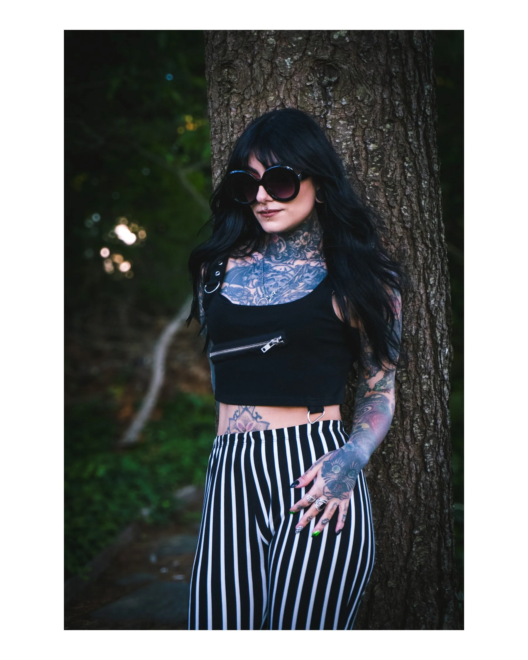 A woman with black hair, wearing large round sunglasses, tattoos on her neck and arms, a nose ring, and striped pants stands leaning against a tree in a wooded area.
