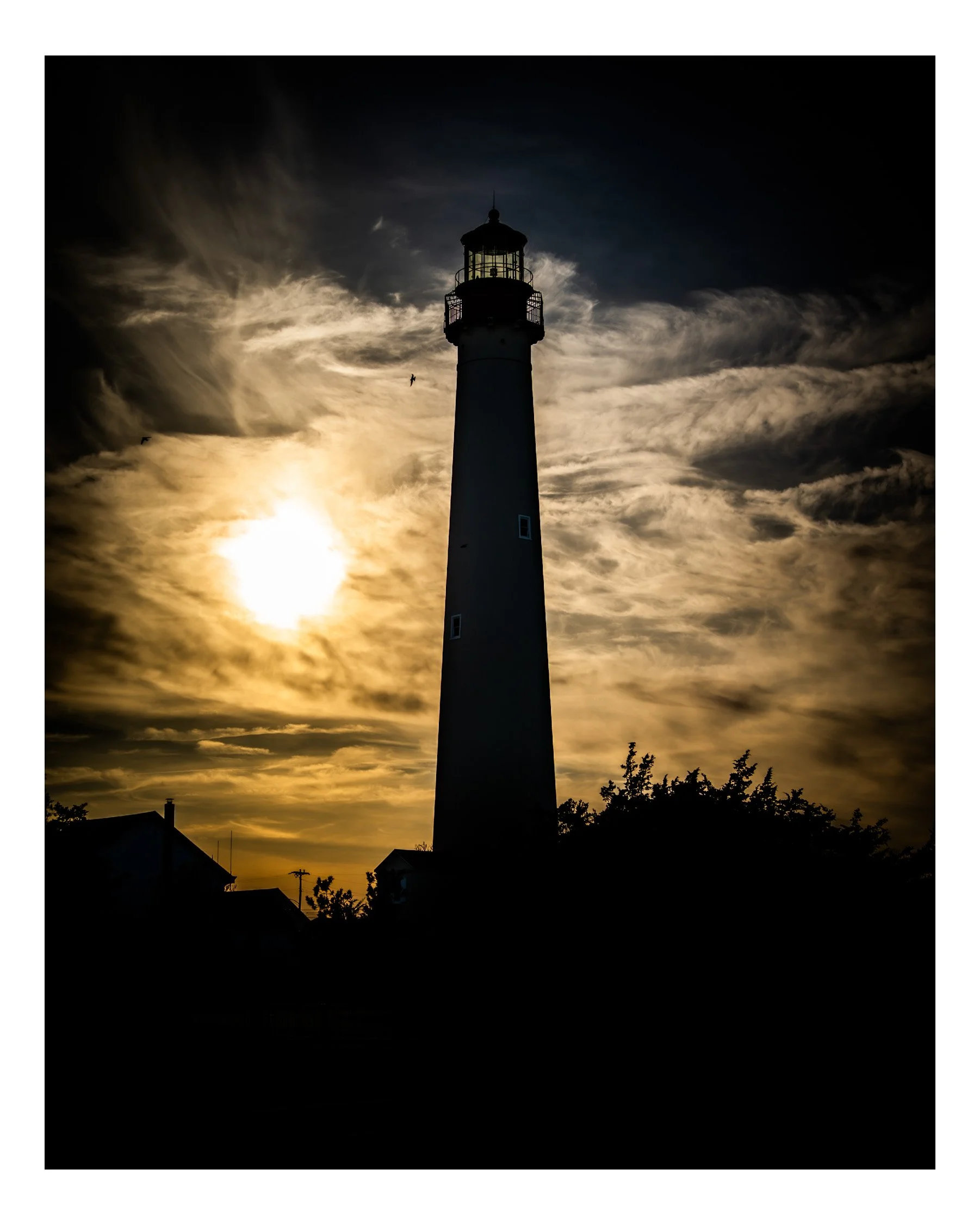Silhouette of a lighthouse against a dramatic sky with clouds and the sun setting or rising, with shadows of houses and trees in the foreground.