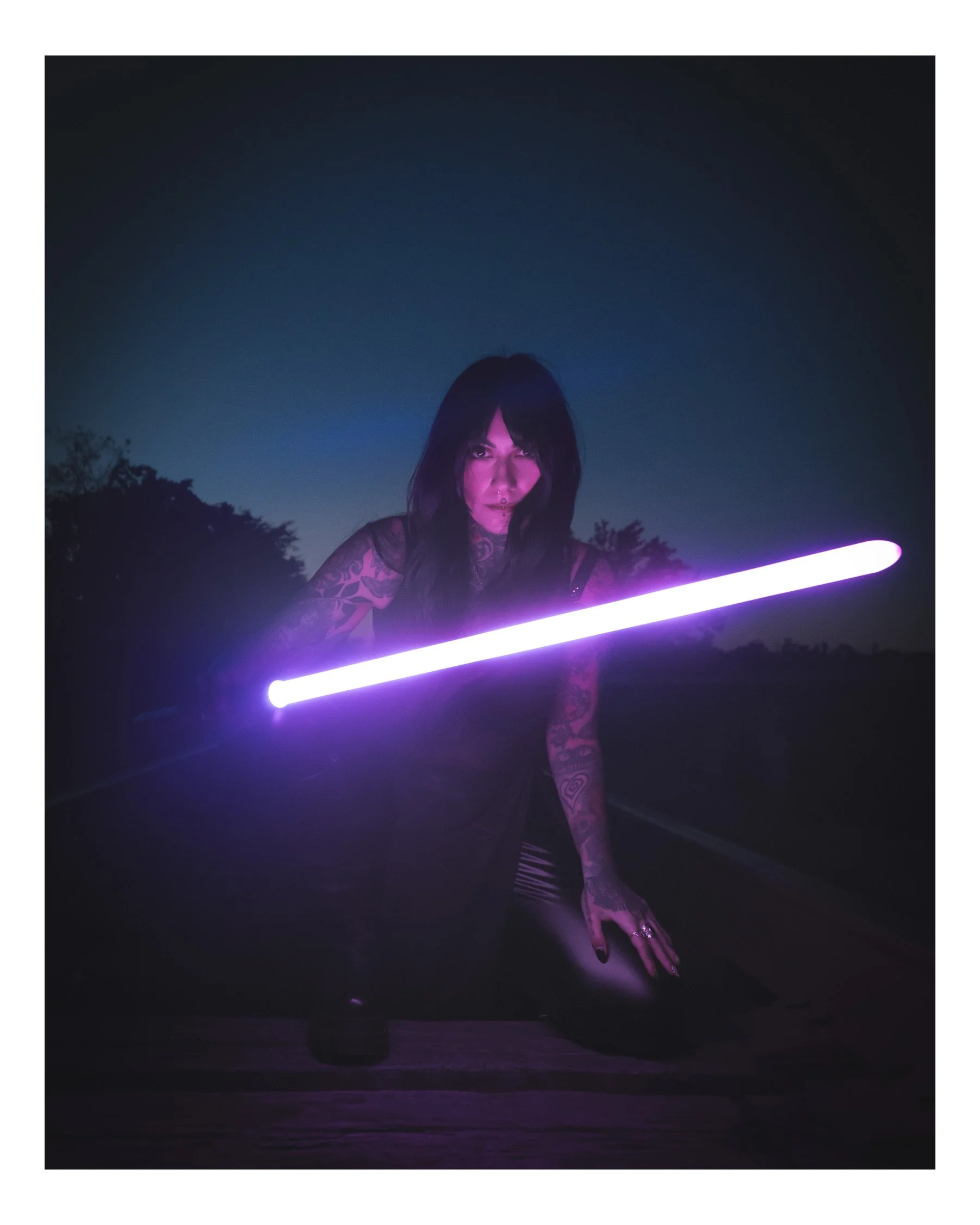 A woman with dark hair and tattoos on her arms is holding a glowing purple lightsaber-like object outdoors at night. The sky is dark with a slight gradient, and trees are visible in the background.
