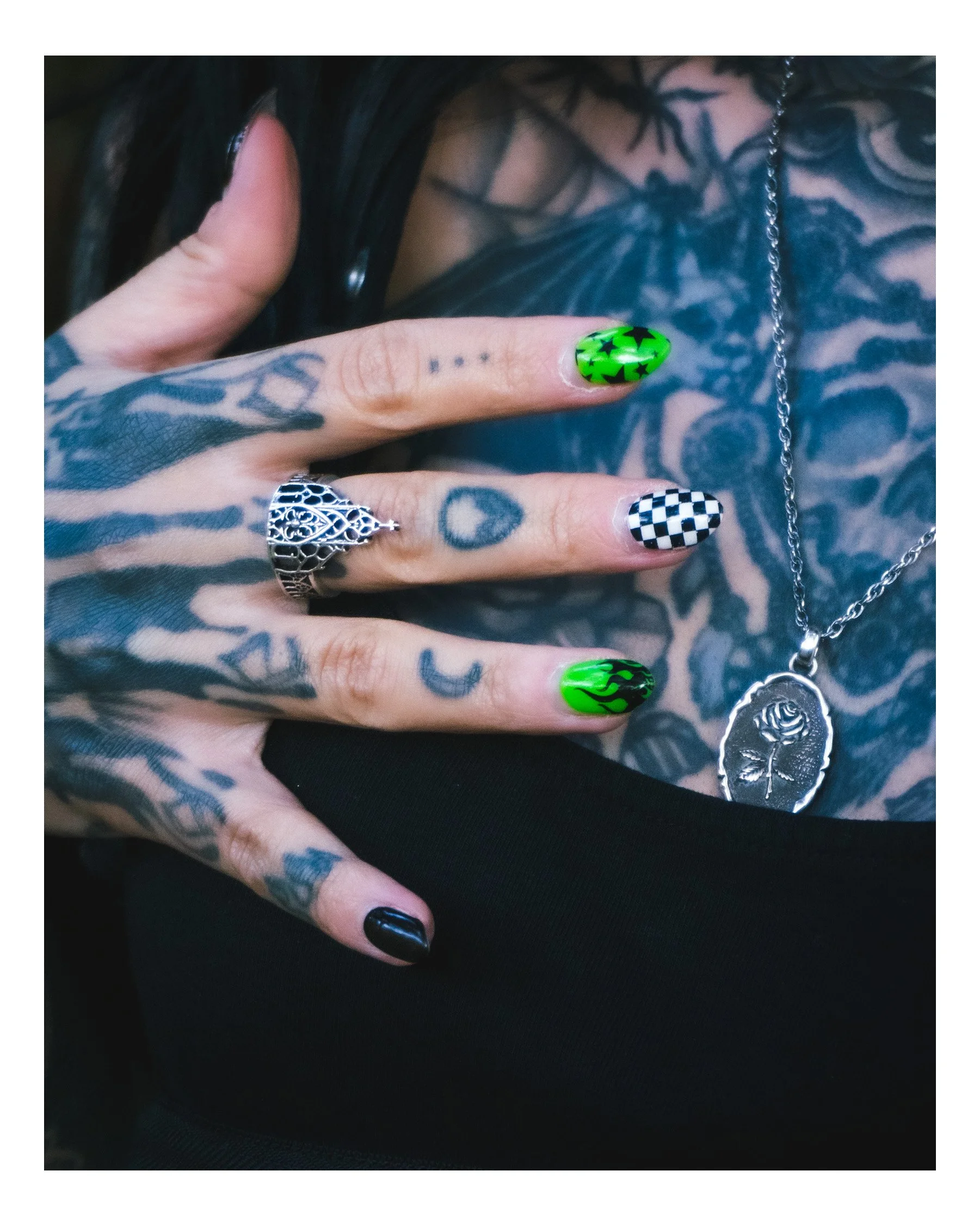 Close-up of a person's hand with tattoos and decorated fingernails, including neon green with black stars, checkered black and white, and black with blue accents, touching or resting on a tattooed chest with a silver chain necklace and black clothing
