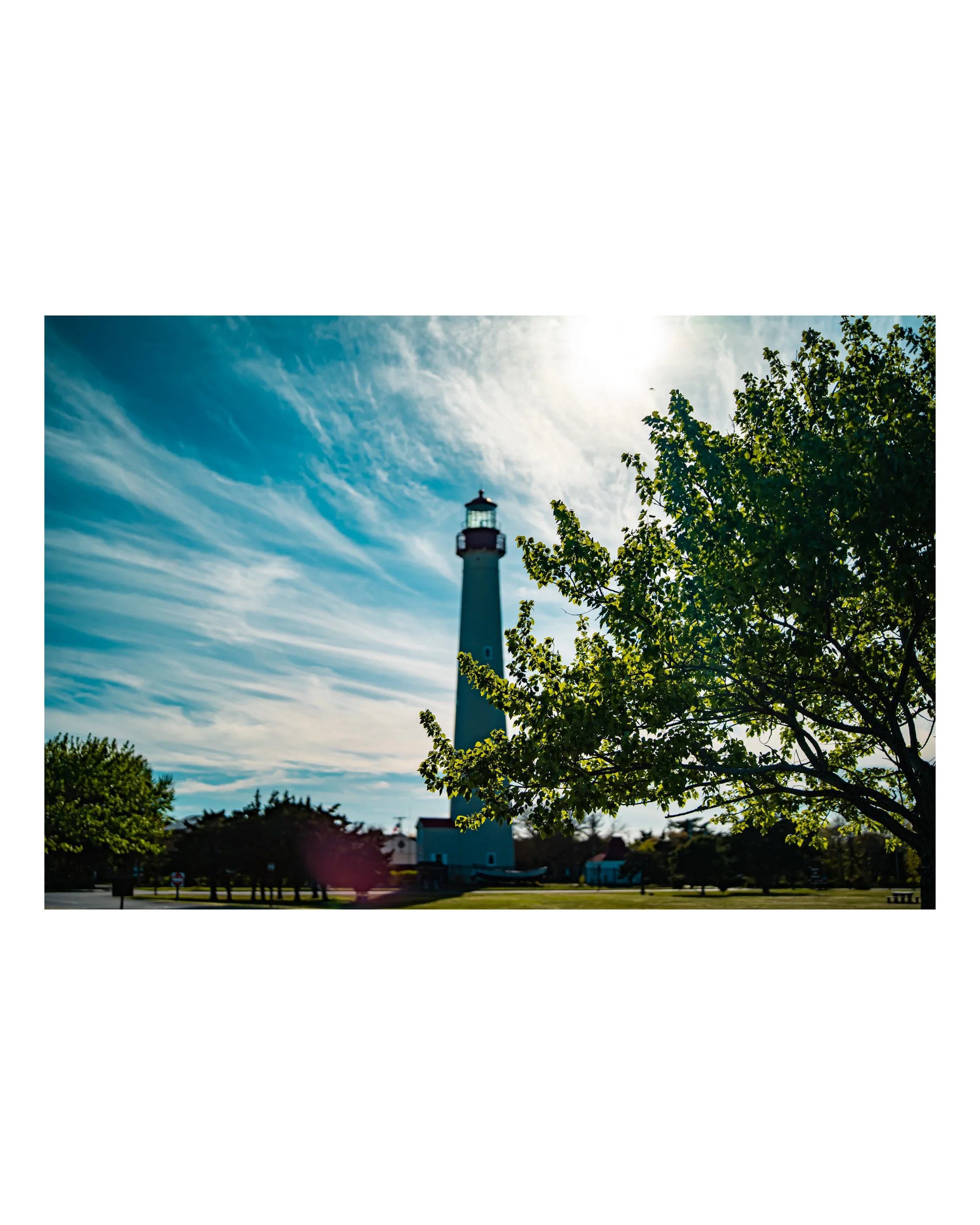 A lighthouse with a dark top and white body in a park with tall trees and a clear blue sky with wispy clouds, illuminated by sunlight.