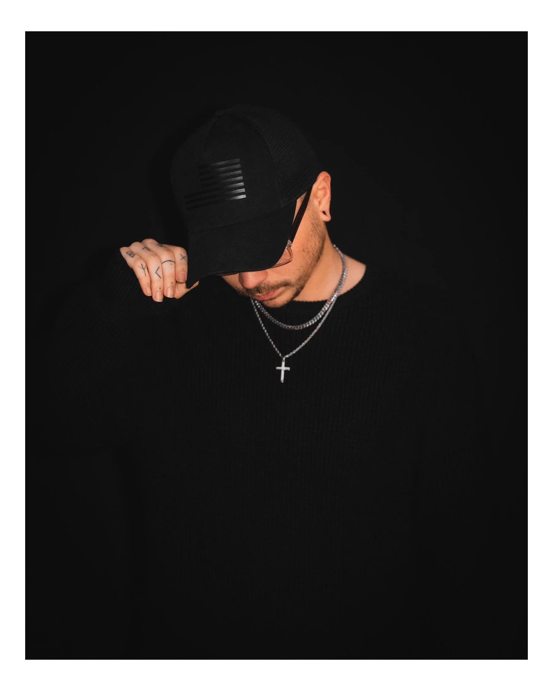 A person in a black cap, glasses, and a black shirt with a cross necklace, against a black background.