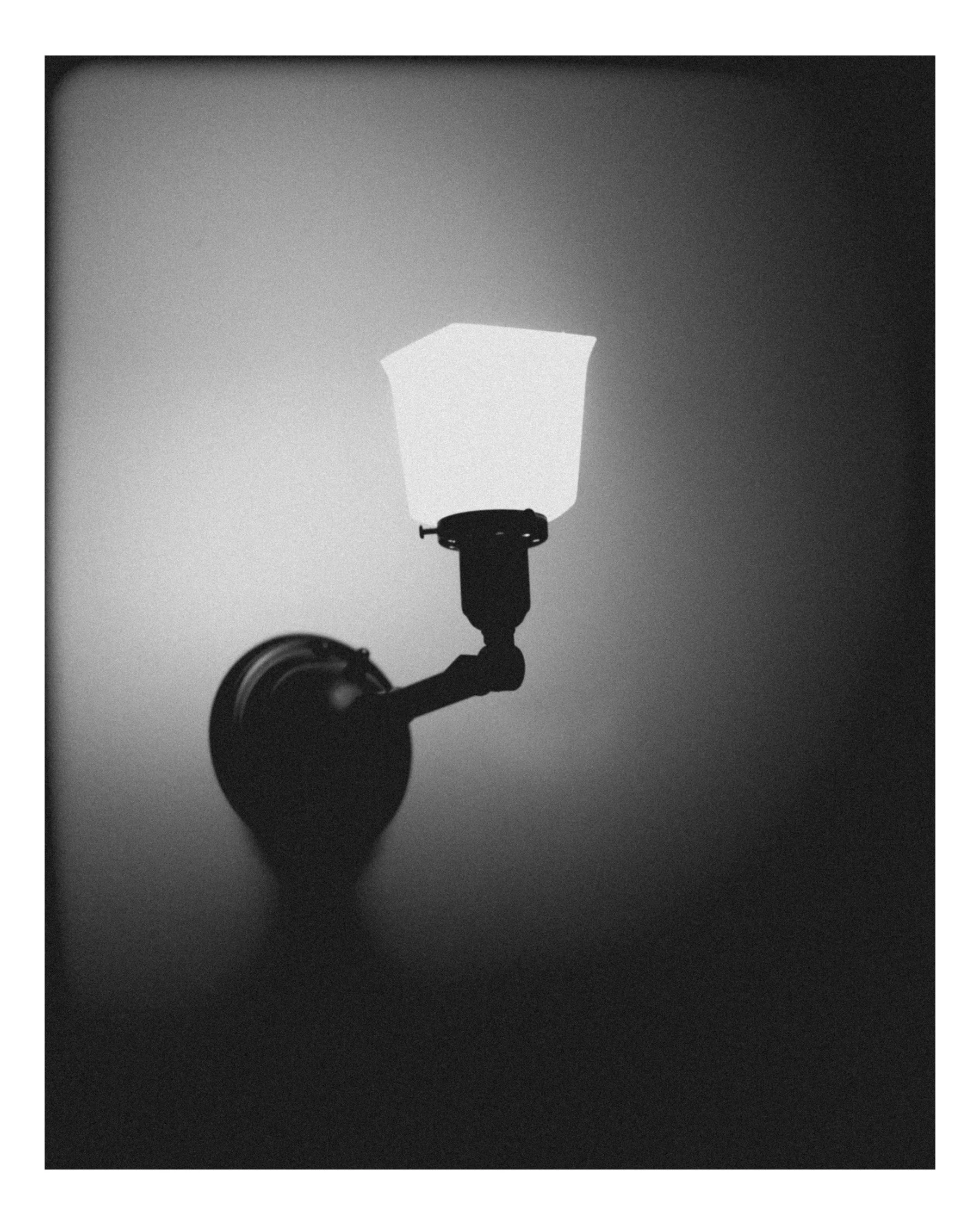 A black and white photo of a street lamp with a hexagonal glass cover, casting a shadow on the wall behind it.