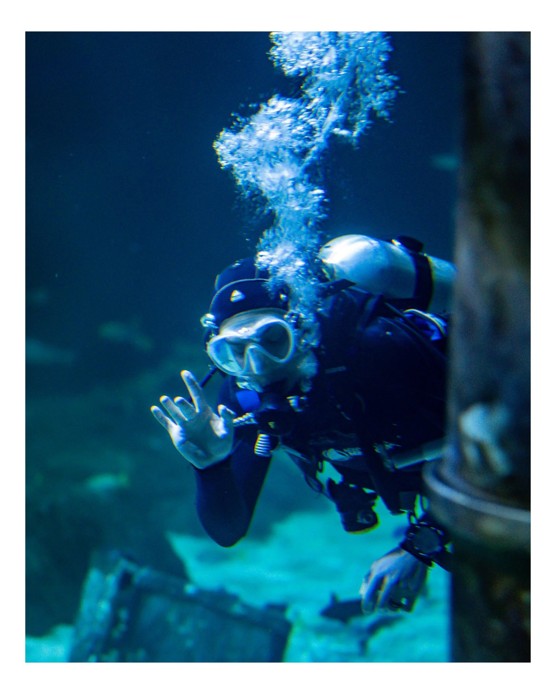 A scuba diver underwater, seen from a side angle, exhaling bubbles into the water with the ocean background visible.