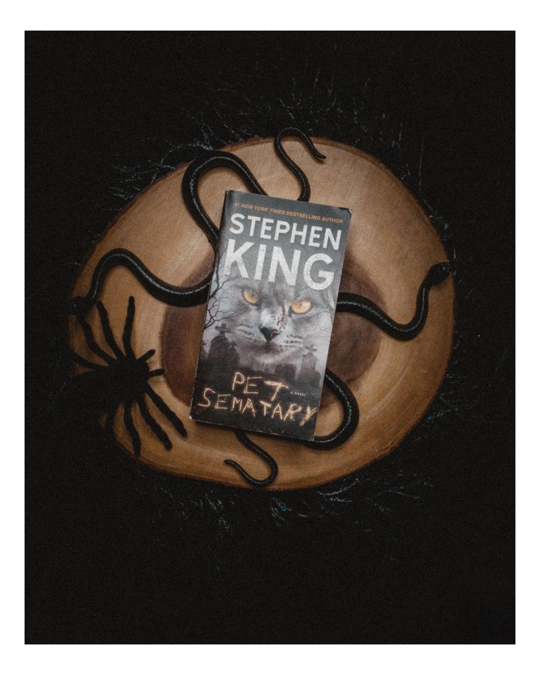 A black spider-shaped jewelry piece holding a book titled "Pet Sematary" by Stephen King, resting on a round wooden surface with a black background.