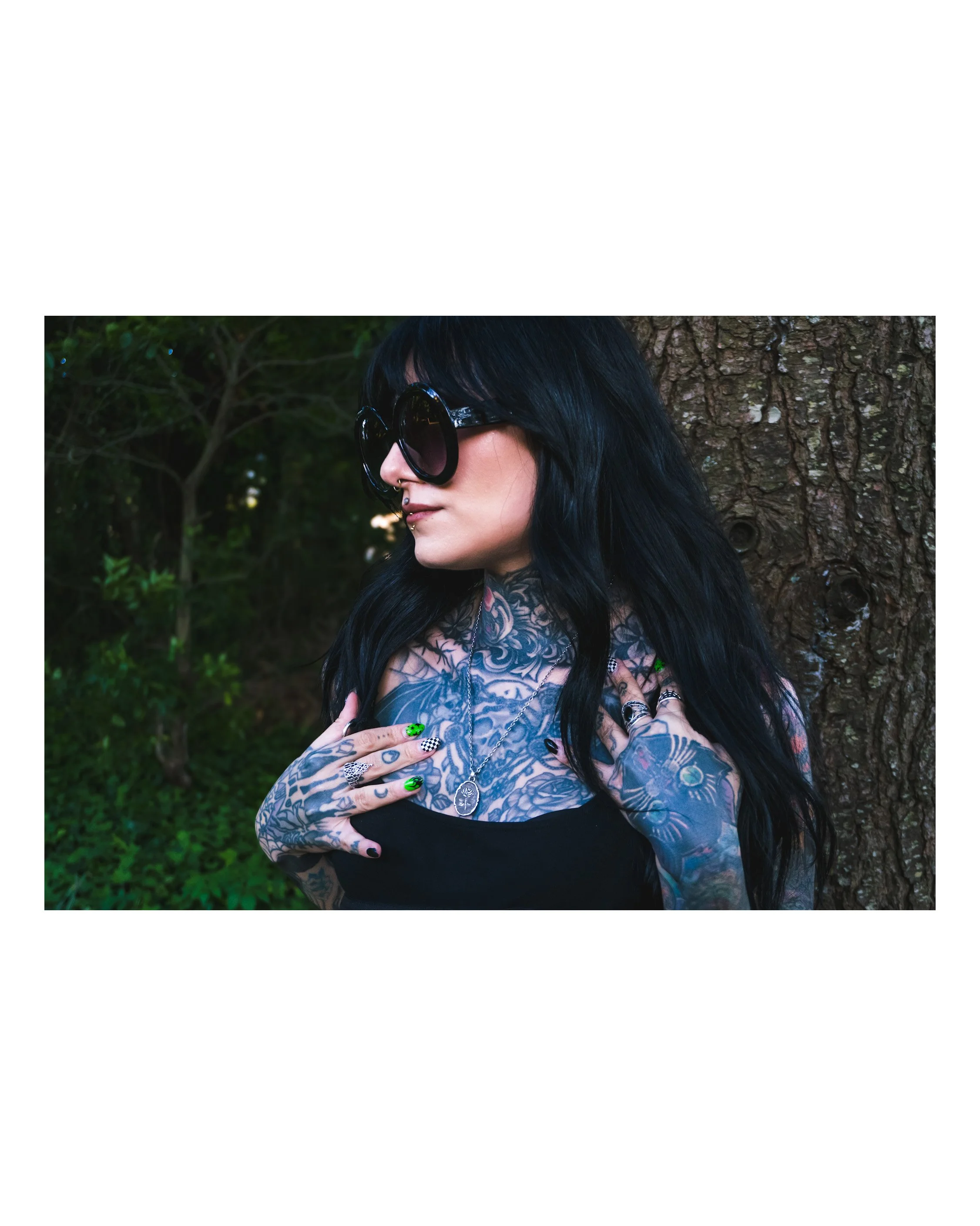 A woman with long black hair and tattoos on her arms and chest wearing large black sunglasses and multiple necklaces, standing outdoors in front of a tree with a textured bark.