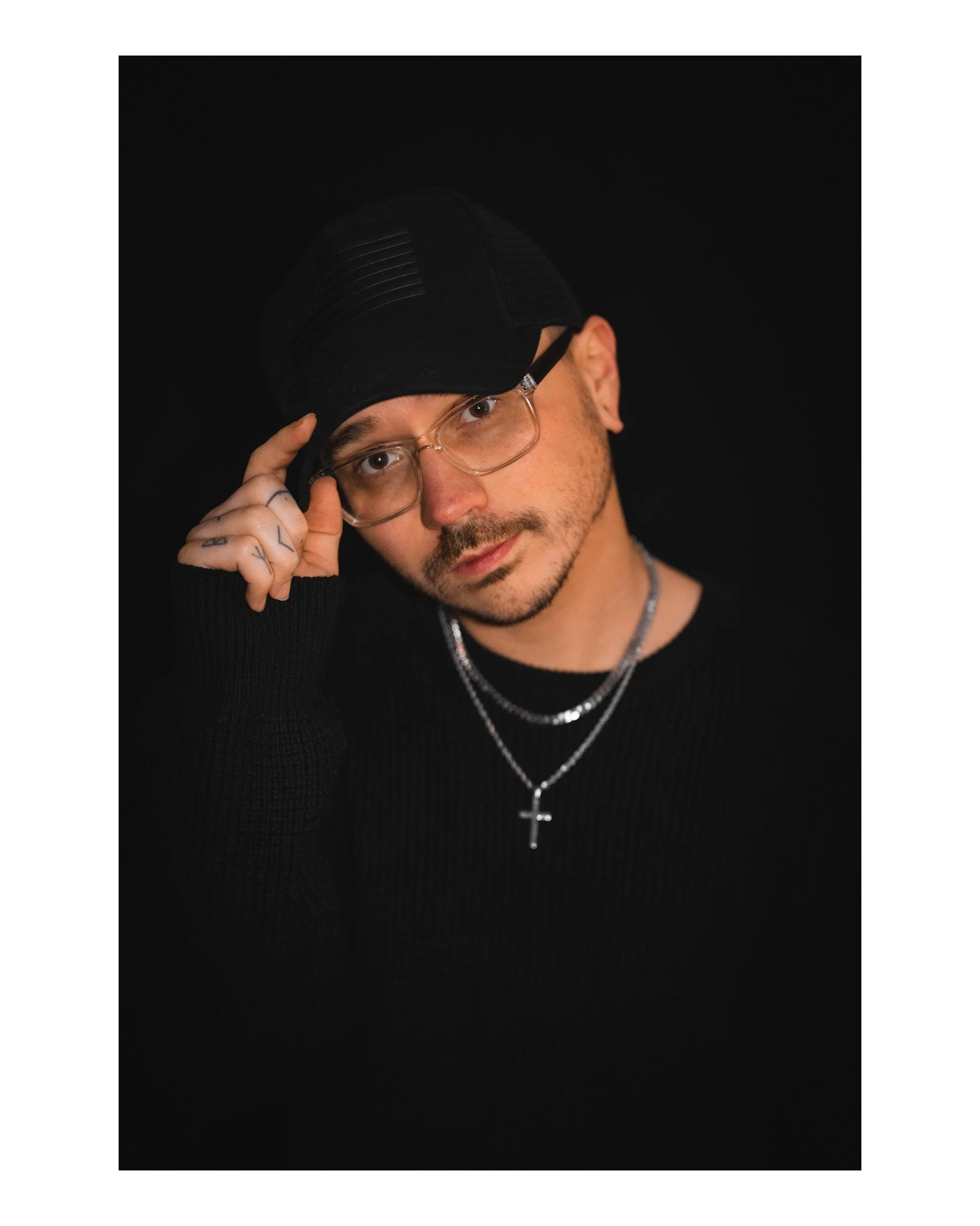 Close-up of a man with facial hair and glasses, wearing a black cap and black clothing, against a black background, adjusting his glasses with his right hand.