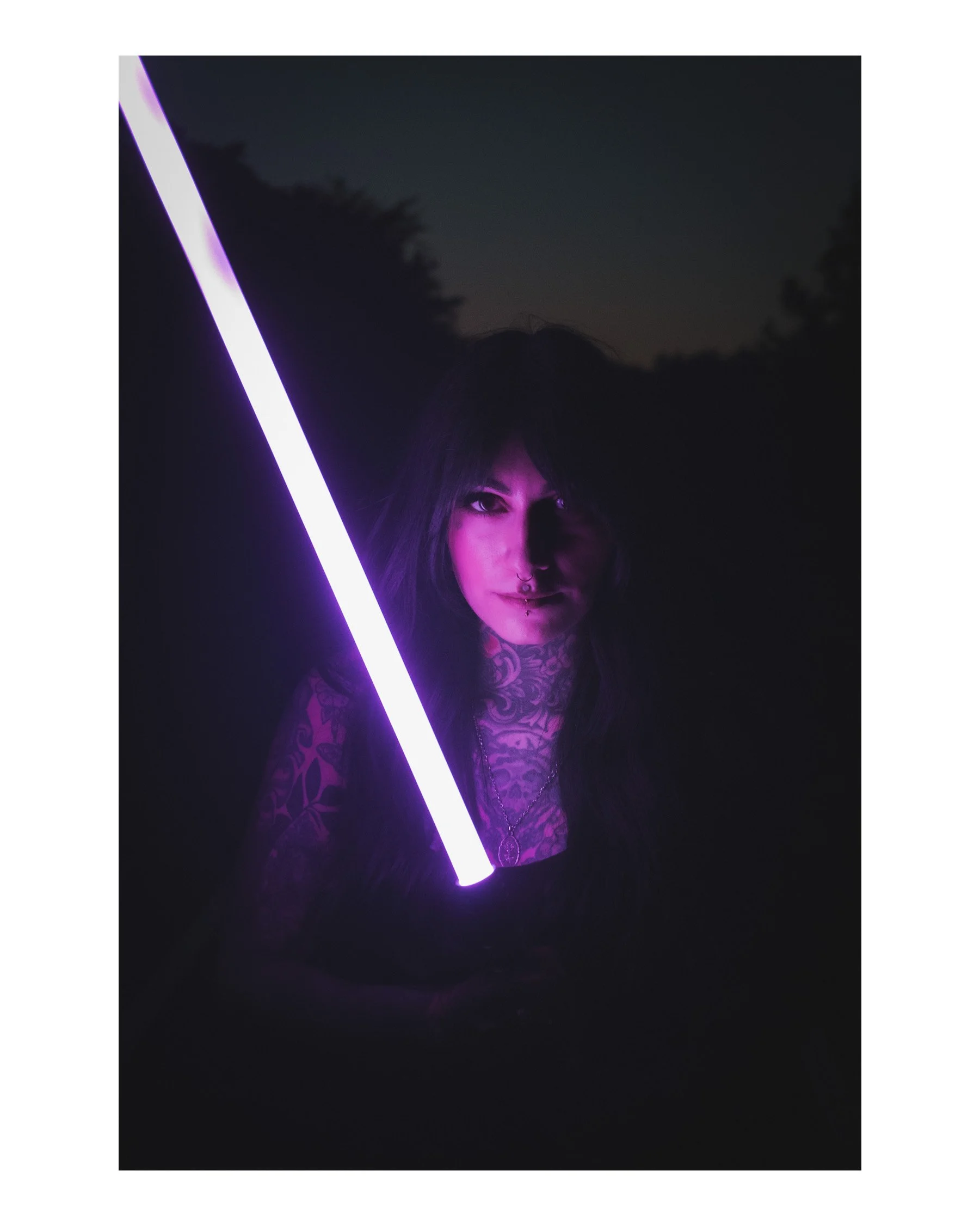 A person with dark hair, tattoos on their chest, and piercing eyes holding a glowing purple light tube in a dark outdoor setting at night.