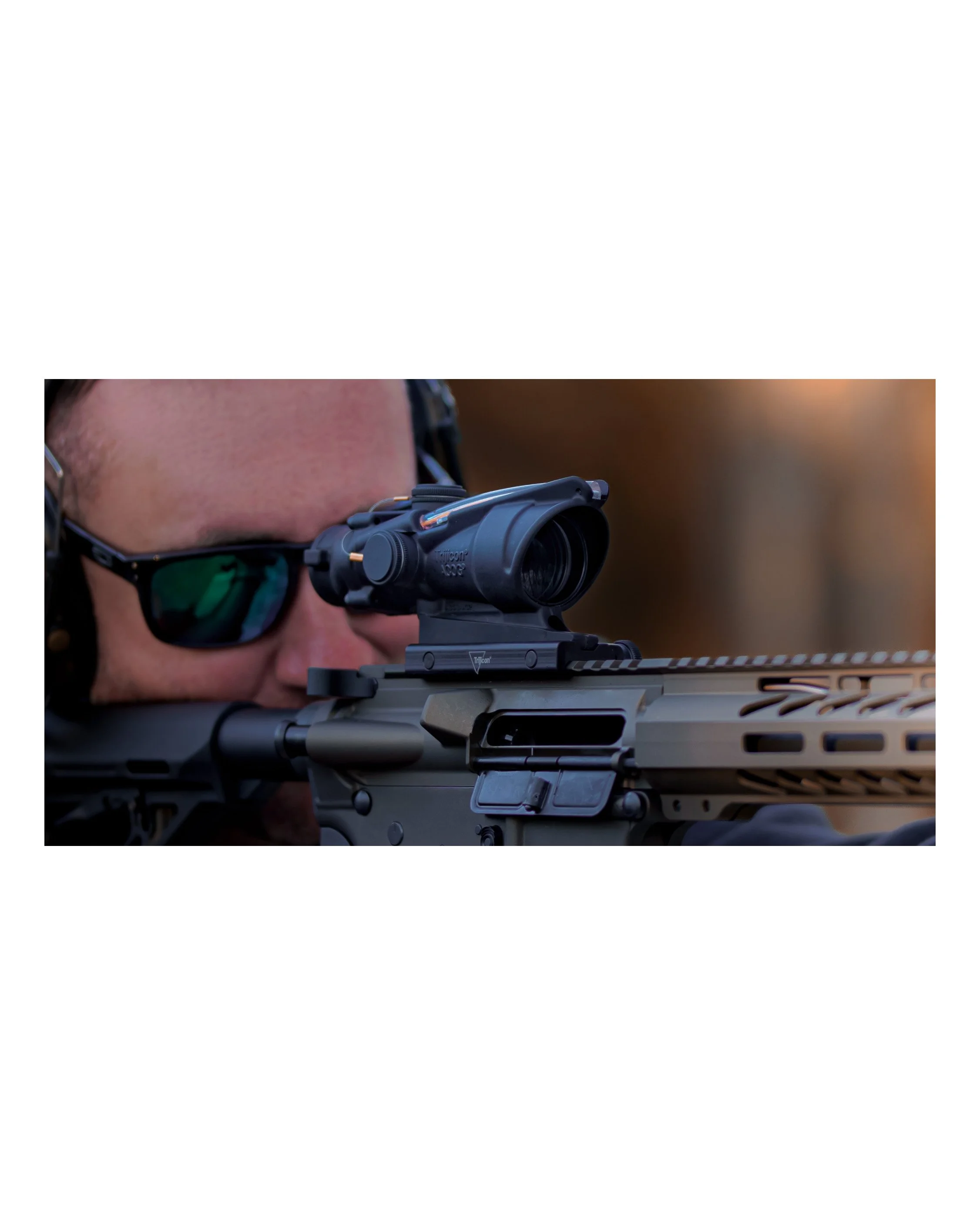 Close-up of a person aiming through a tactical rifle scope, wearing sunglasses and hearing protection.