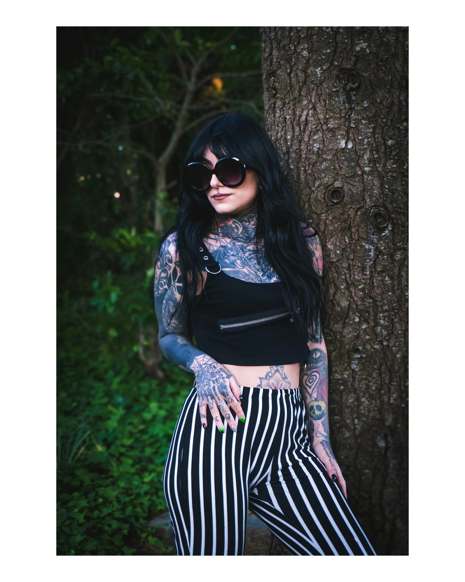 A woman with black hair and sunglasses standing outdoors next to a tree, wearing tattoos, a black crop top, and striped pants.