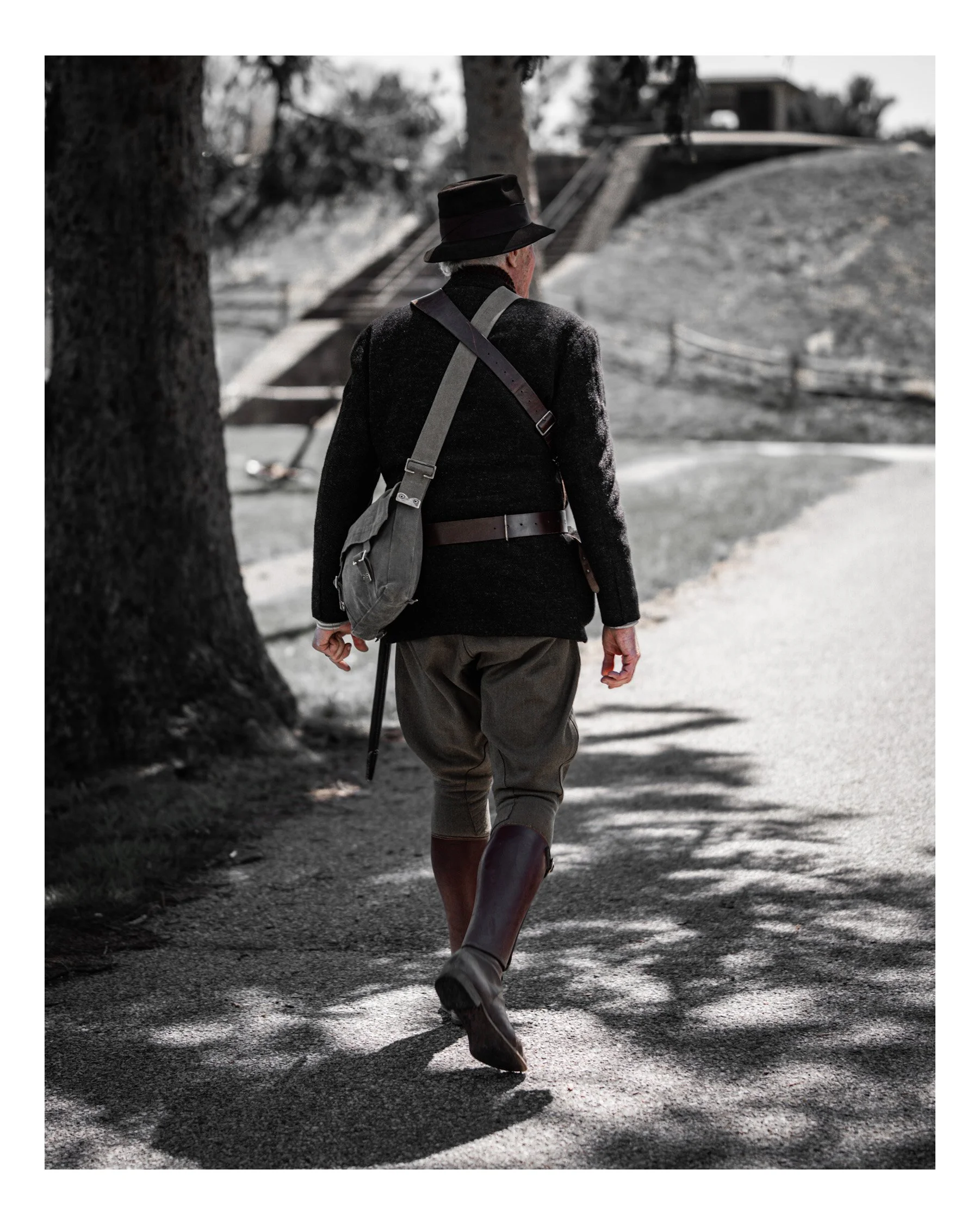 A man dressed as a World War I soldier walking outdoors near a large tree, with a silhouette of a building in the background.