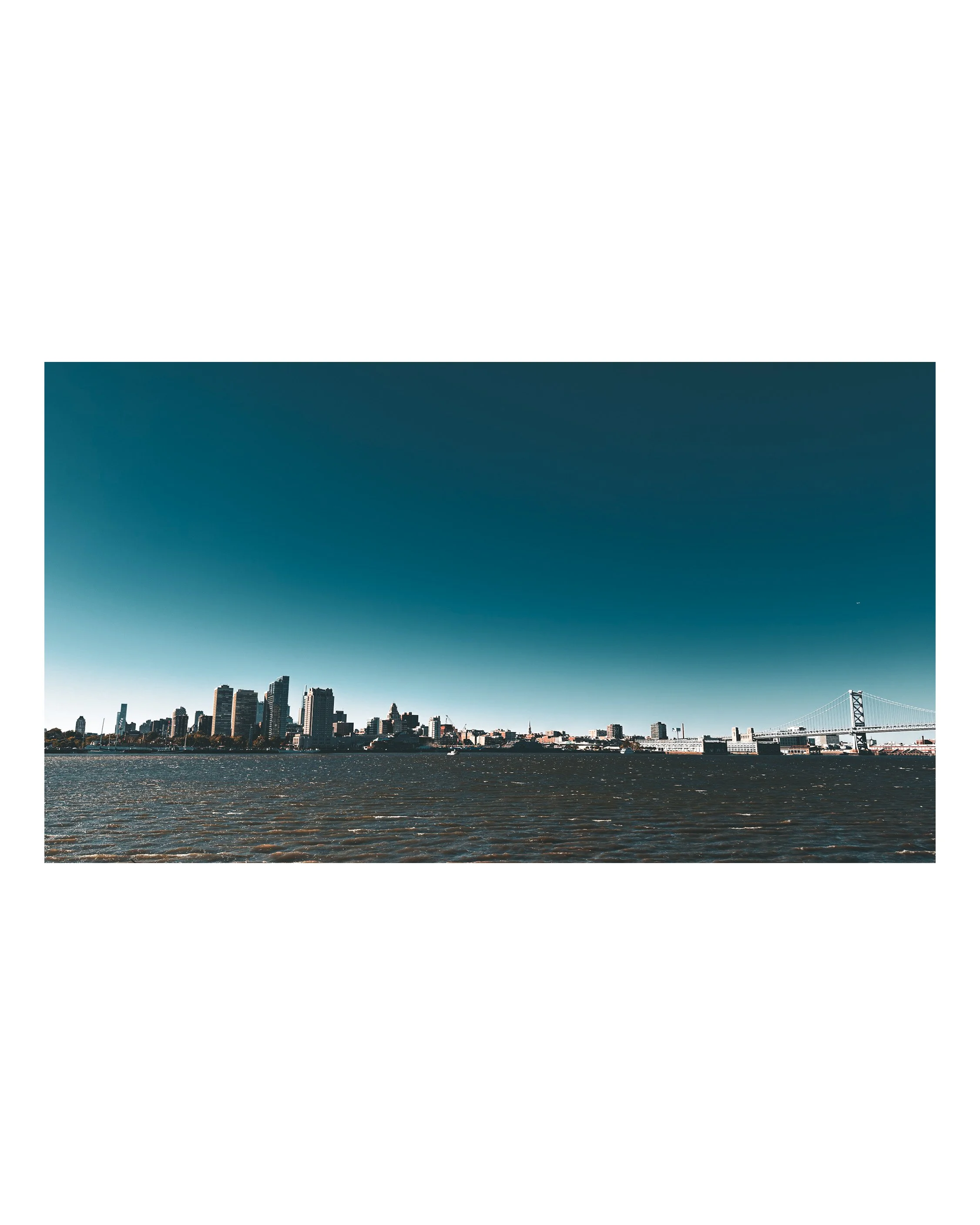 City skyline seen from across a body of water with a bridge on the right. The sky is partly cloudy with a gradient of dark to light blue.