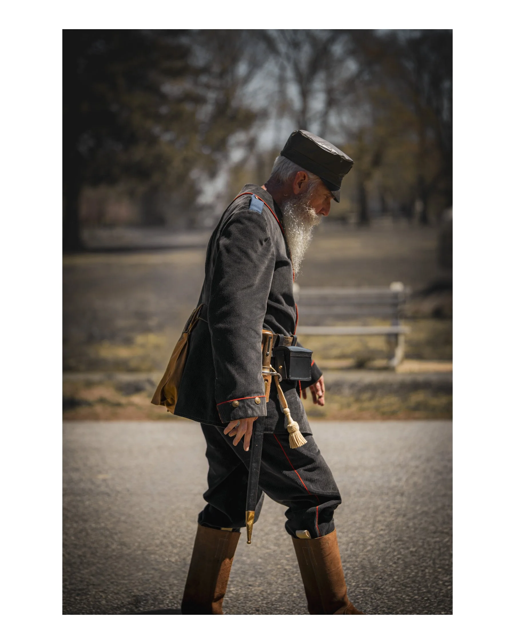 A man dressed in historical military uniform, wearing tall leather boots, a gray jacket with red piping, and a black hat with a flat top, standing outdoors during daytime with trees and a cloudy sky in the background.