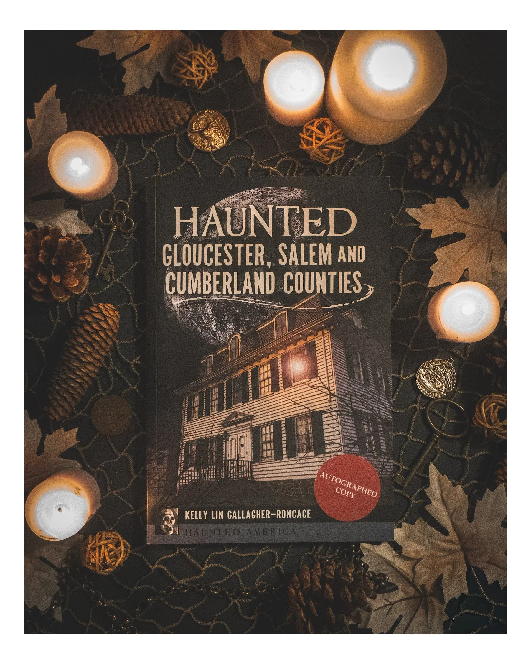 Book titled 'Haunted America' by Kelly Lin Gallagher-Roncace surrounded by candles, pine cones, leaves, decorative spheres, and keys on a dark tablecloth.