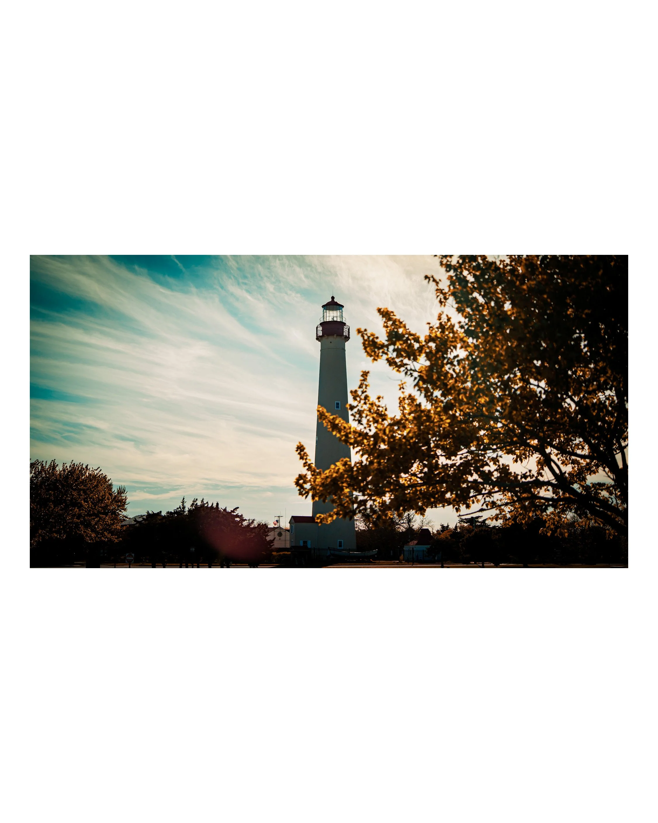 Lighthouse standing tall against a background of a sky with wispy clouds, with trees in the foreground and some small buildings at the base.